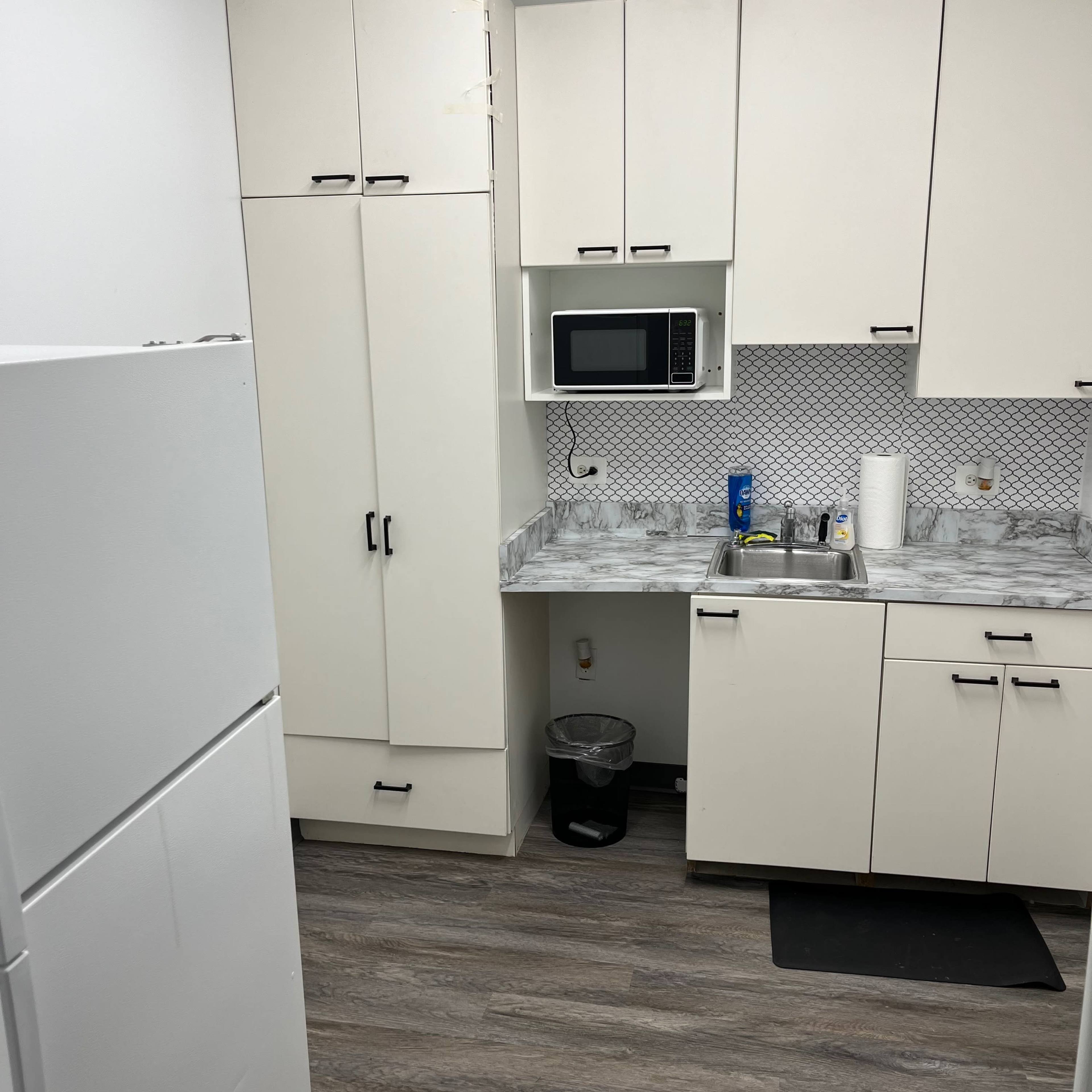 A small kitchen area with white cabinets, a sink, a microwave, and a refrigerator, set against gray laminate flooring.