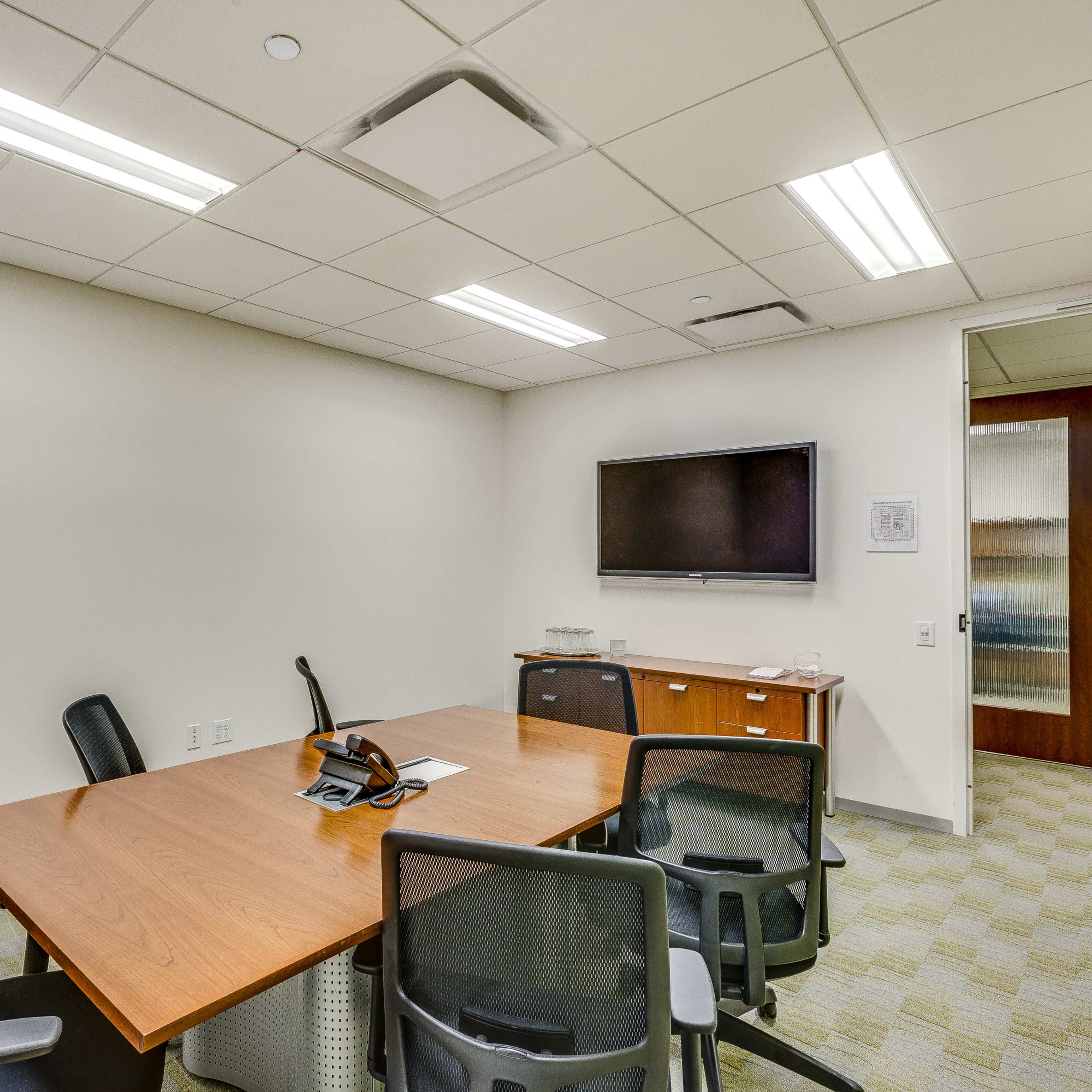 A small meeting room features a wooden table surrounded by black chairs, with a flat-screen TV mounted on the wall and a wooden cabinet in the corner.