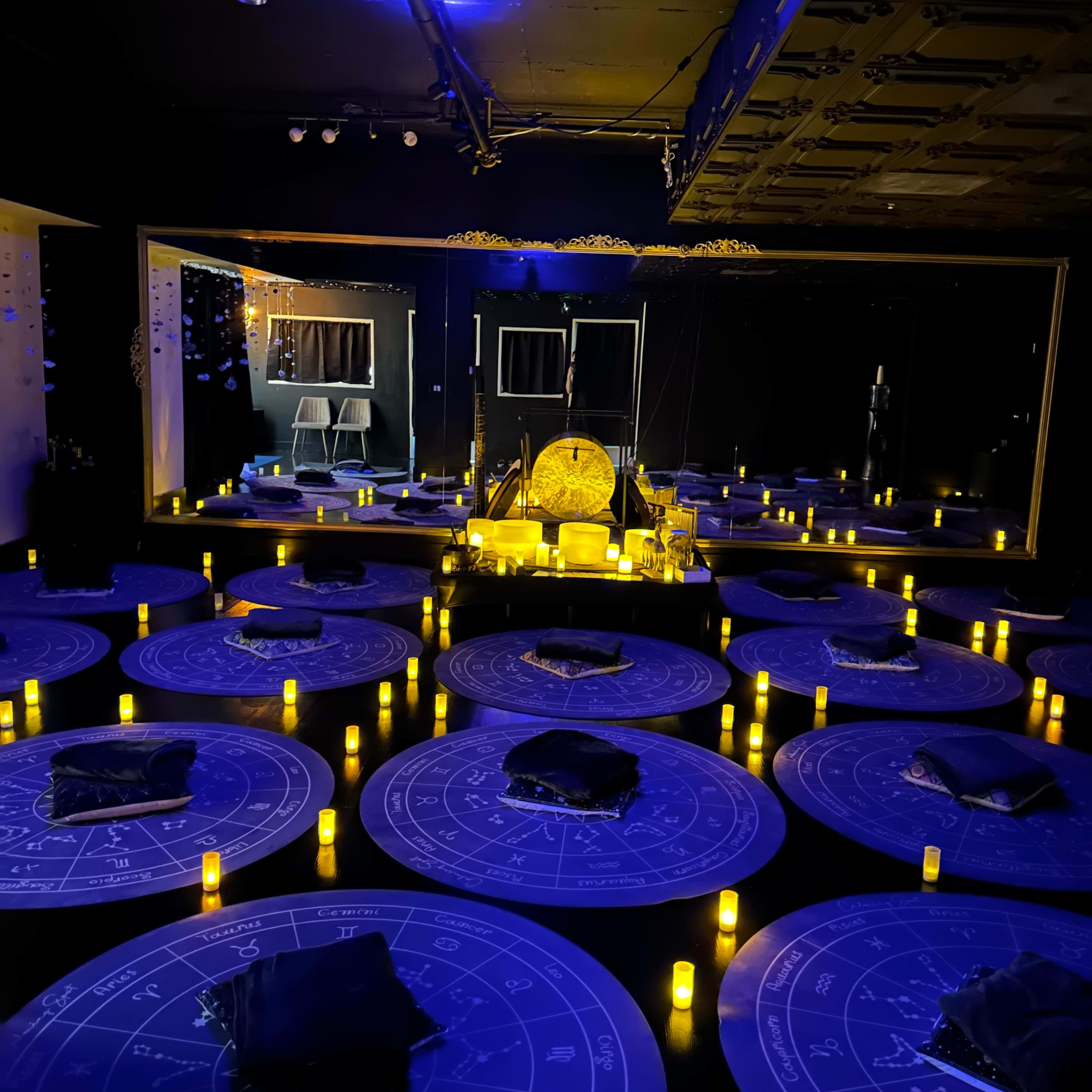 A dimly lit room decorated with circular tables, each featuring astrological symbols, surrounded by flickering candles and a central illuminated area with an ornate object.