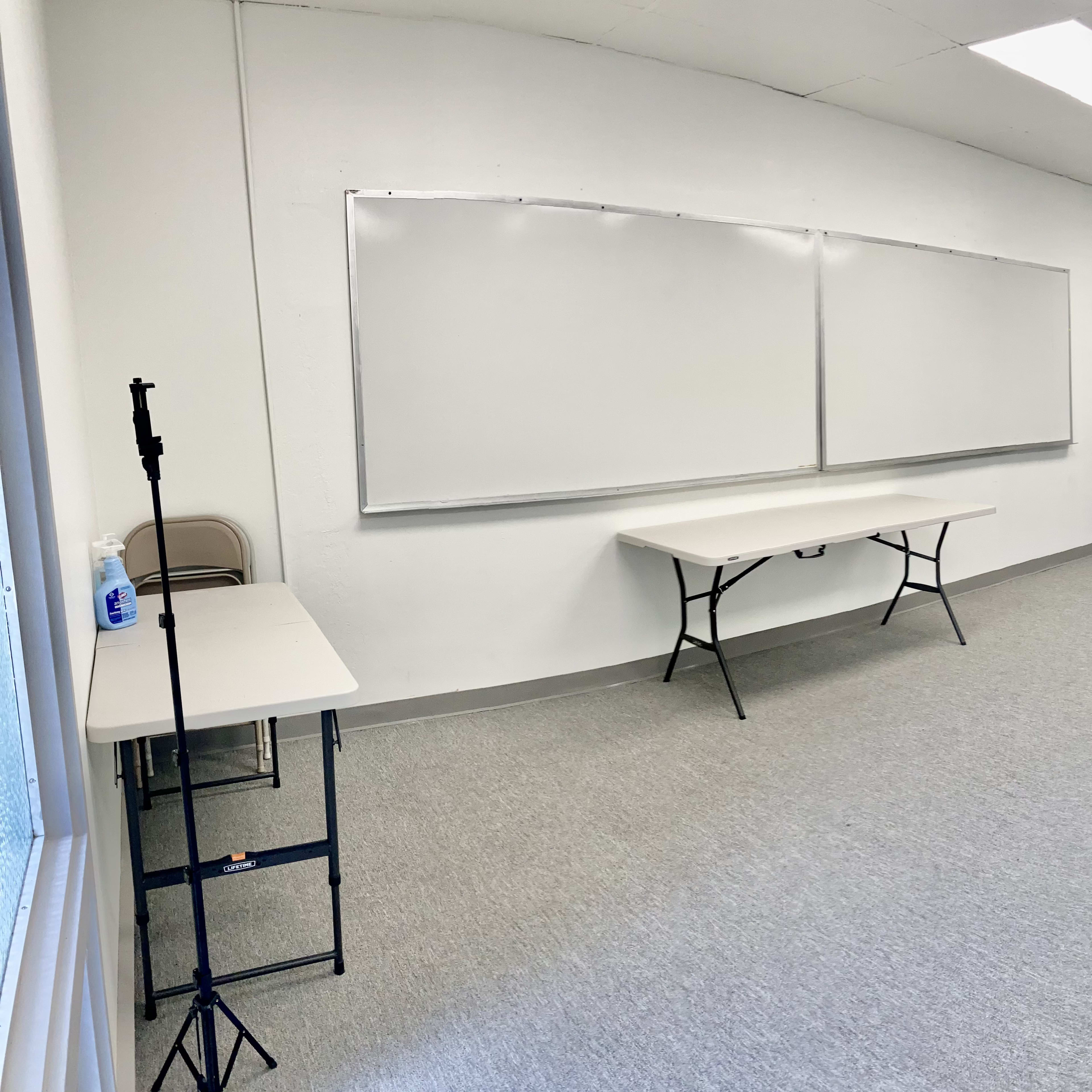 A spacious, empty classroom with a gray carpet, two whiteboards, a table, and minimal furniture.