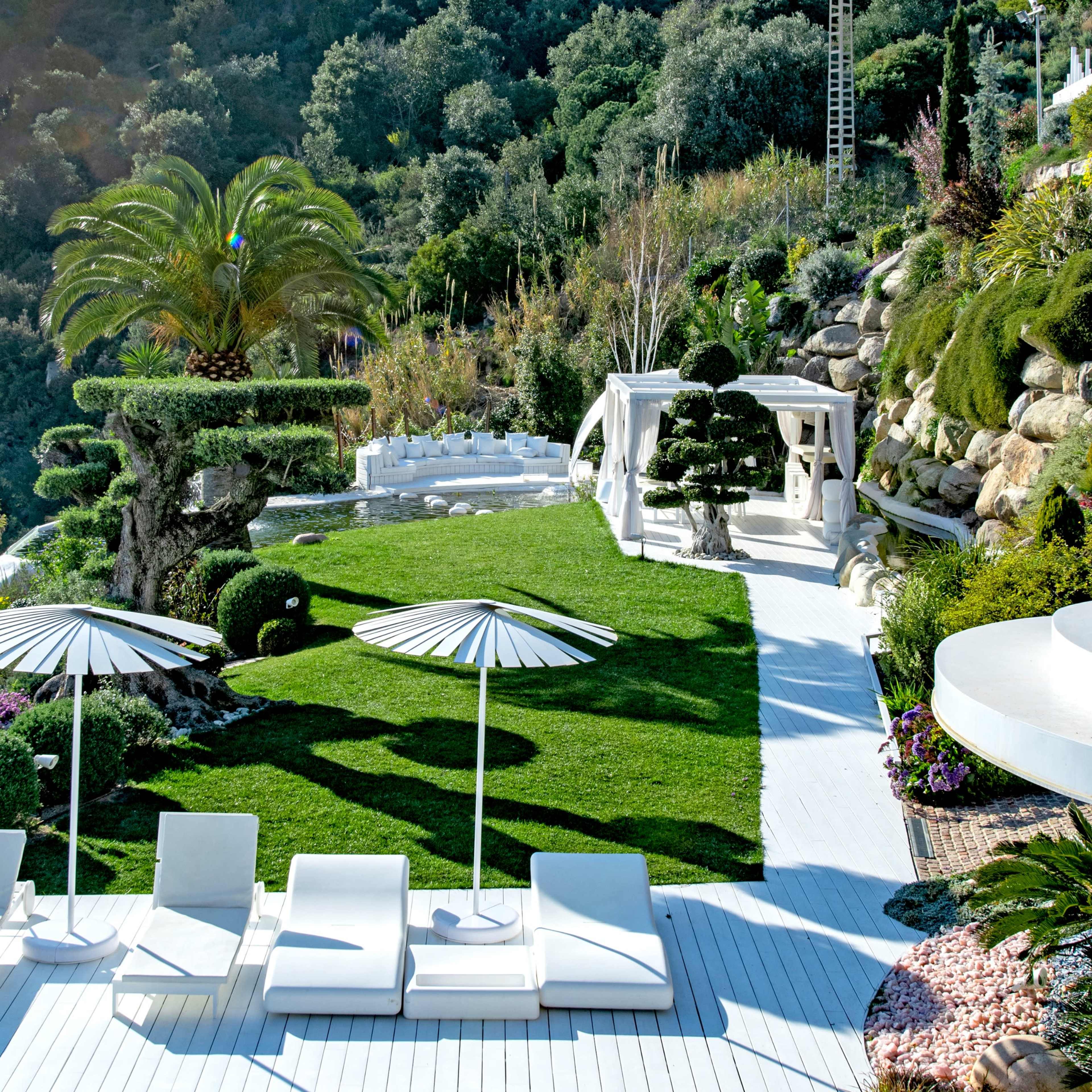 A serene outdoor pool area featuring white lounge chairs, large umbrellas, and carefully manicured gardens with ornamental shrubs and palm trees.