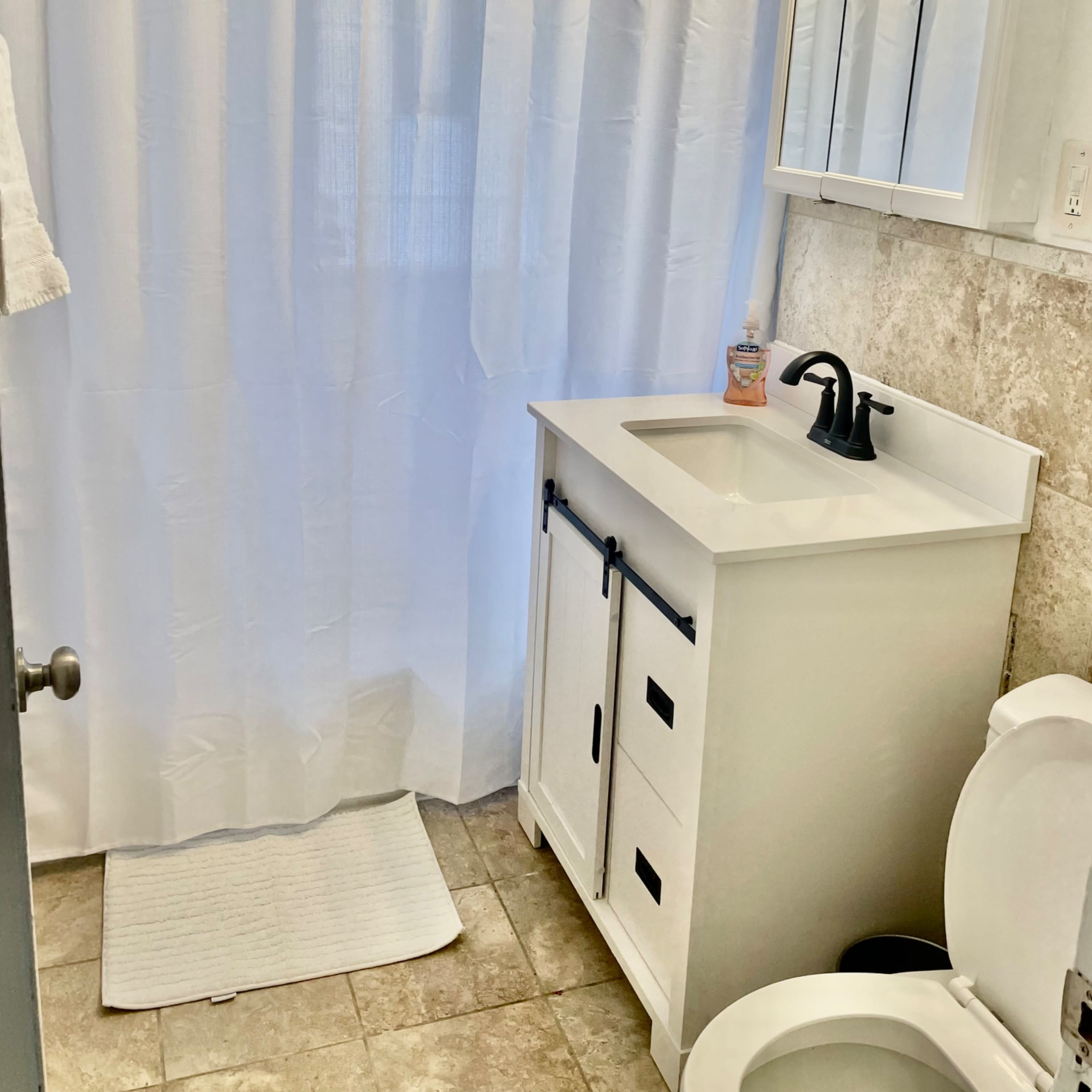 The bathroom features a white shower curtain, a sink with a black faucet, a mirror with light fixtures above, and a toilet.