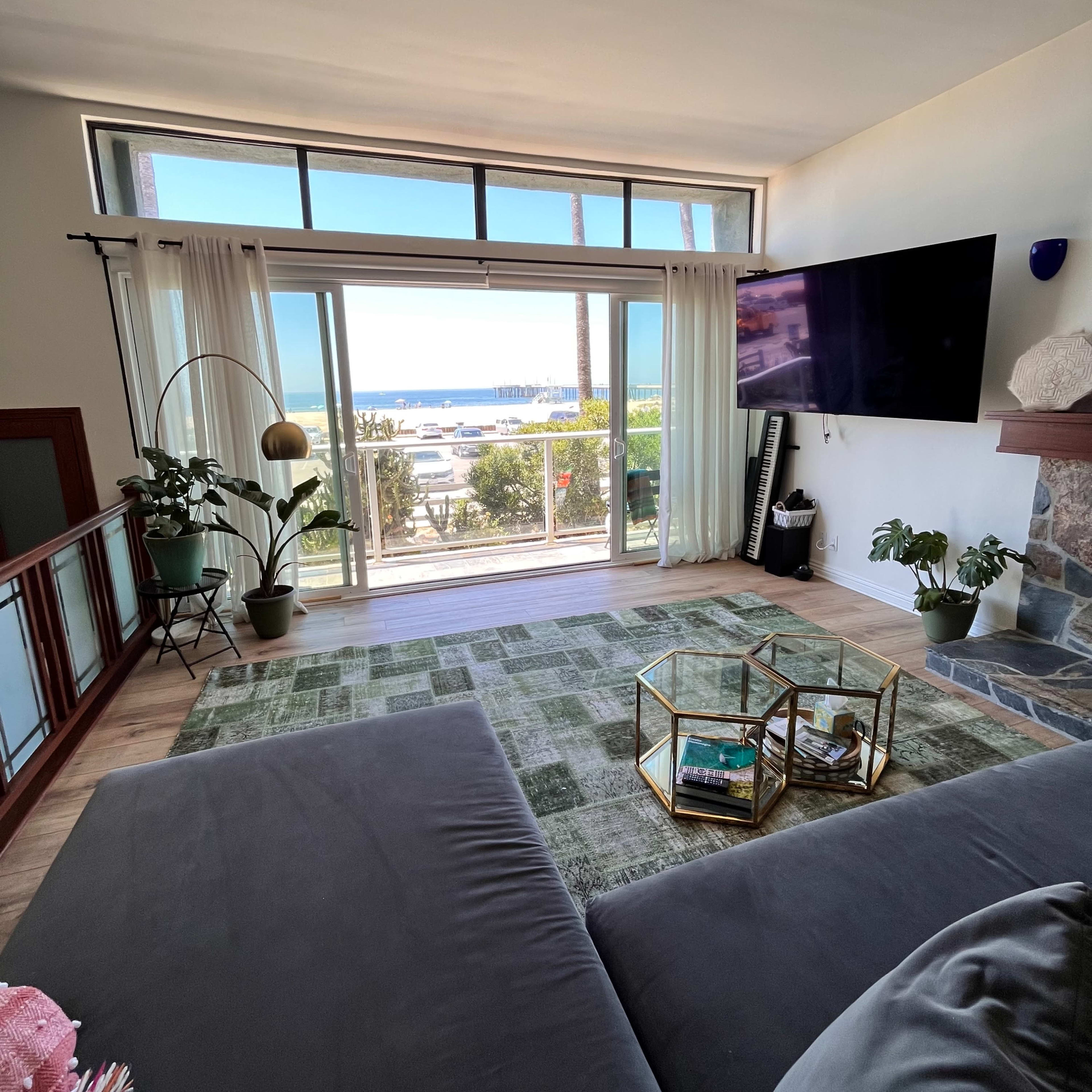 The living room features large windows with a view of the ocean, a gray sofa, a green rug, and a stone fireplace.