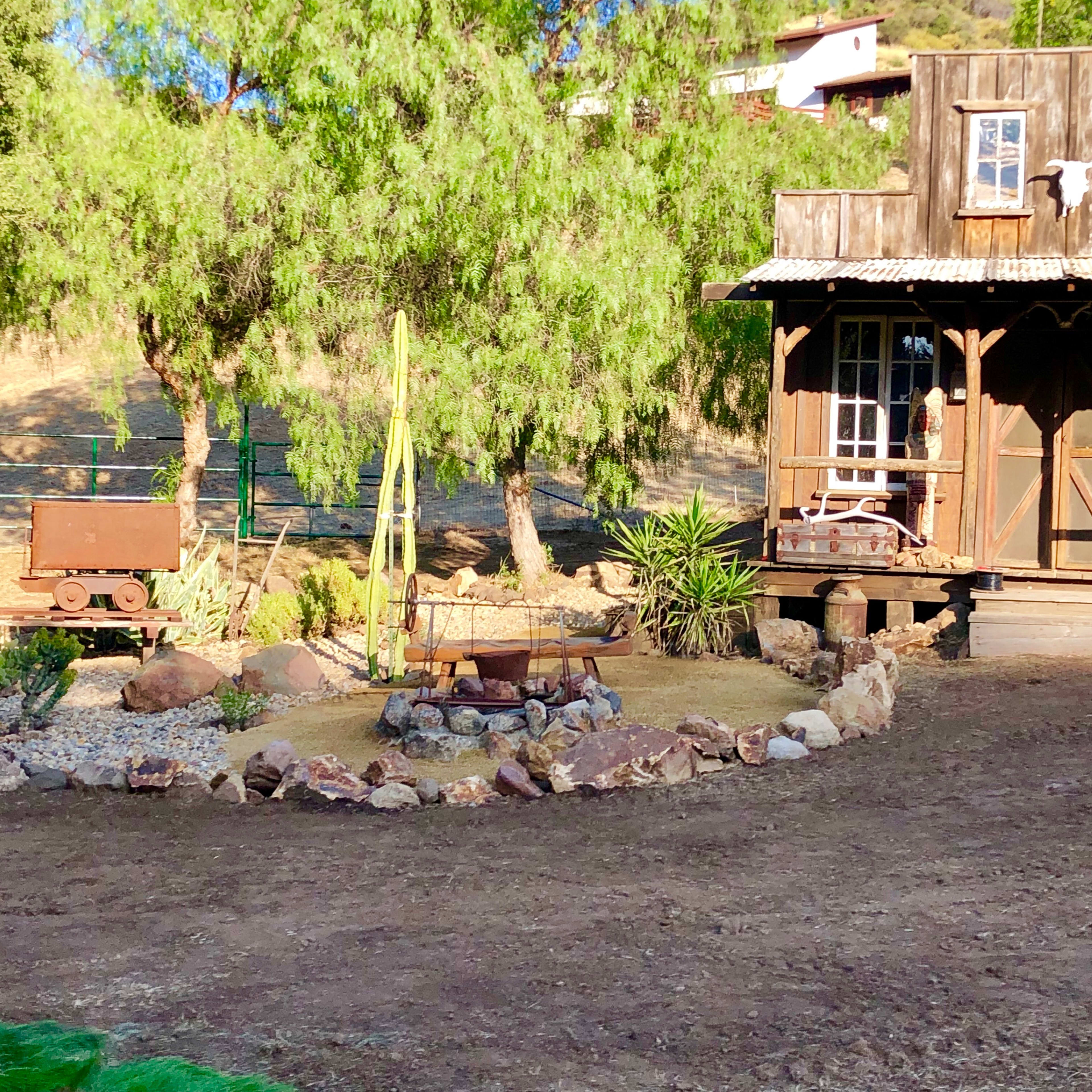 A rustic wooden cabin surrounded by landscaped gravel and stone pathways, with a small wagon and circular fire pit in the foreground.