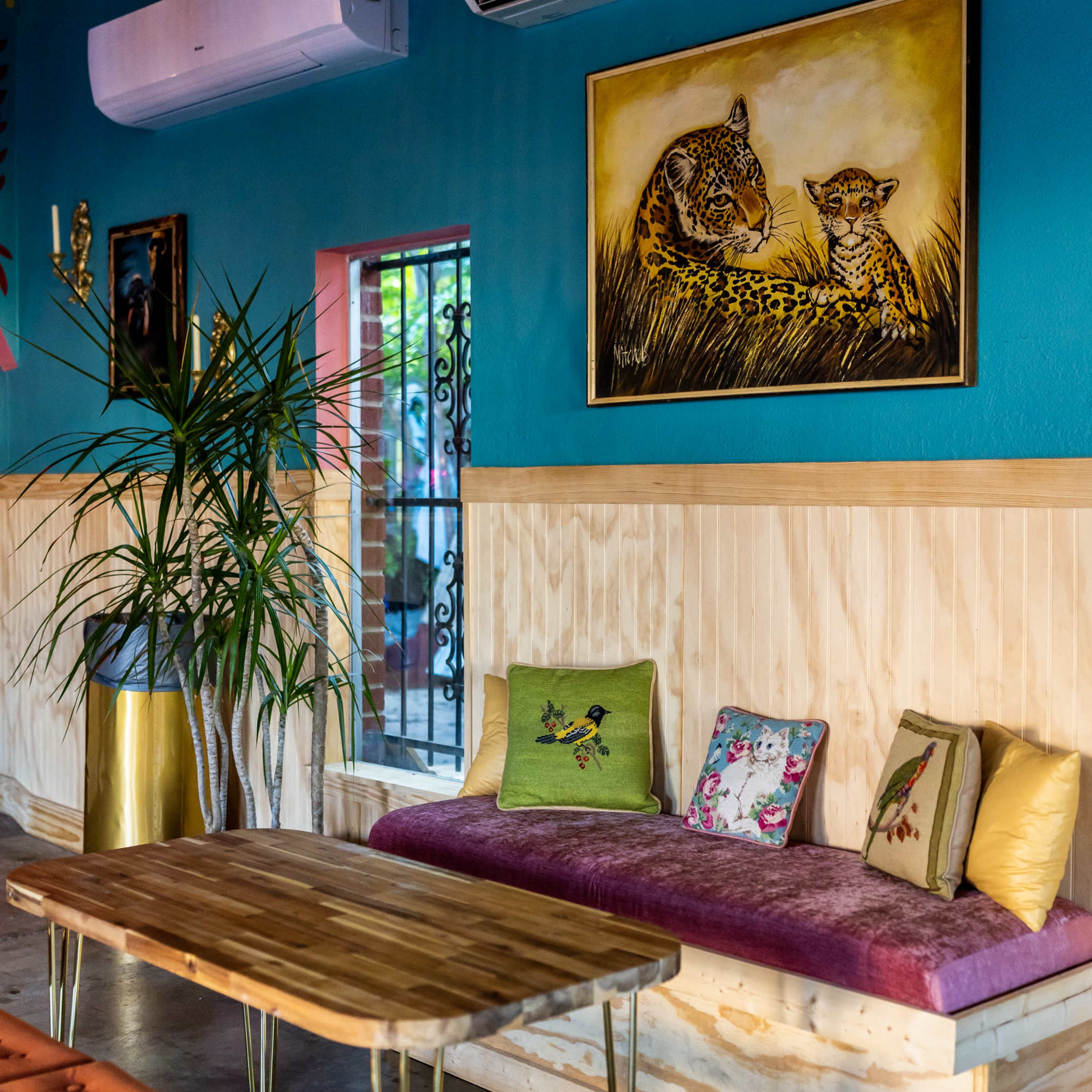 A cozy seating area in a vibrant room, featuring a wooden bench with colorful cushions, a rustic table, and air conditioning units mounted on the blue walls.