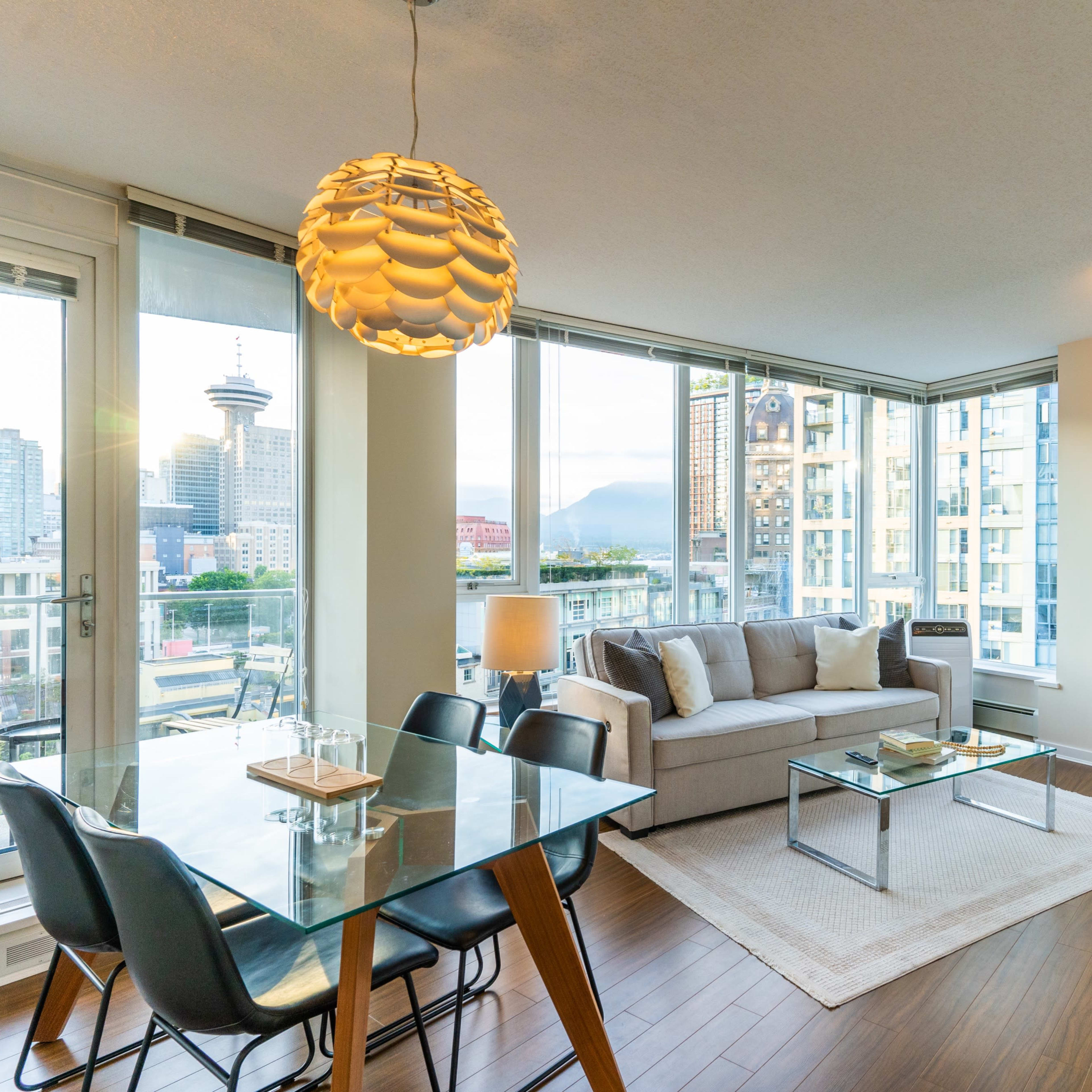 A modern living room and dining area feature large windows with city views, a sofa, a dining table, and a decorative plant.