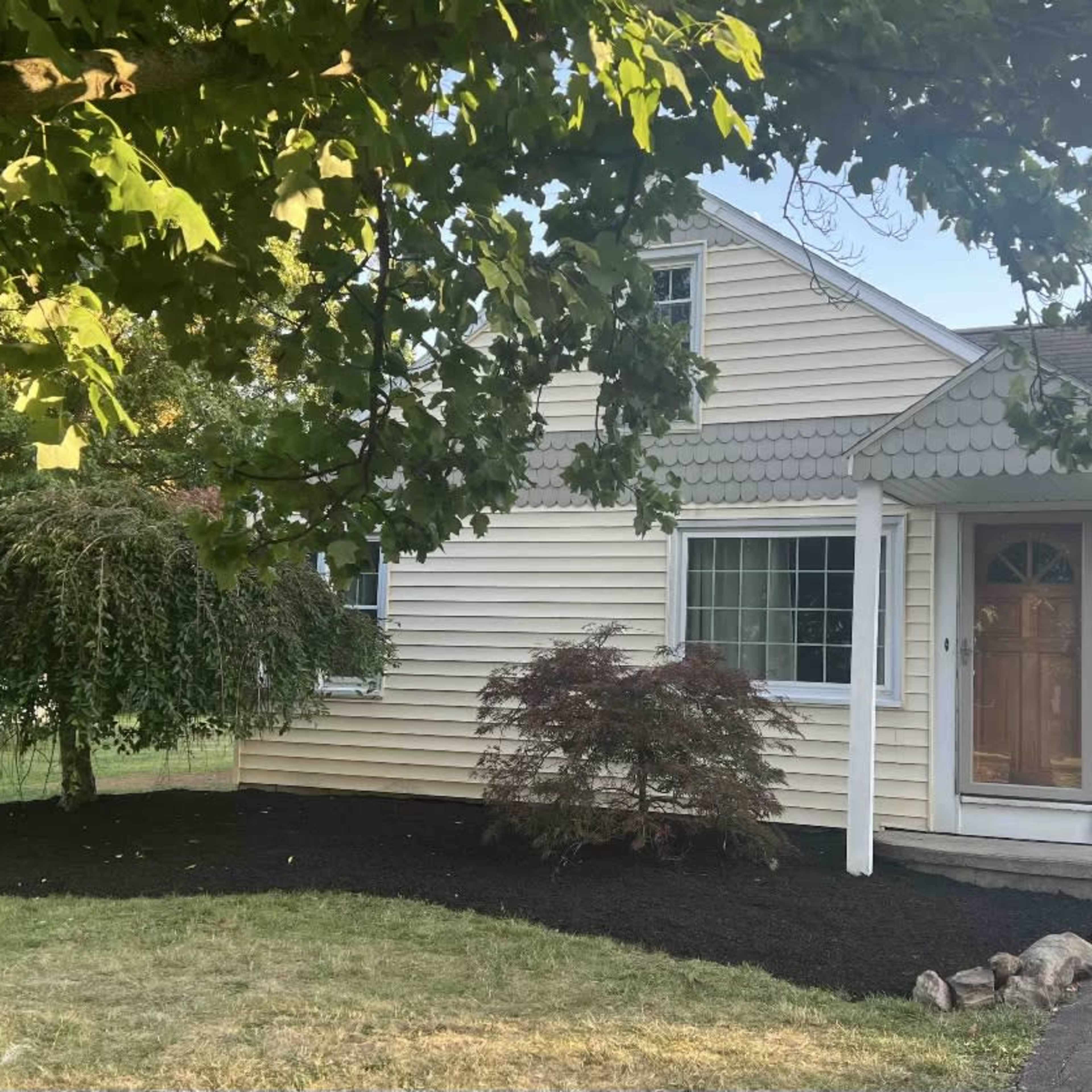 A yellow house with a shingled roof and a front door, surrounded by green trees and freshly mulched flower beds.
