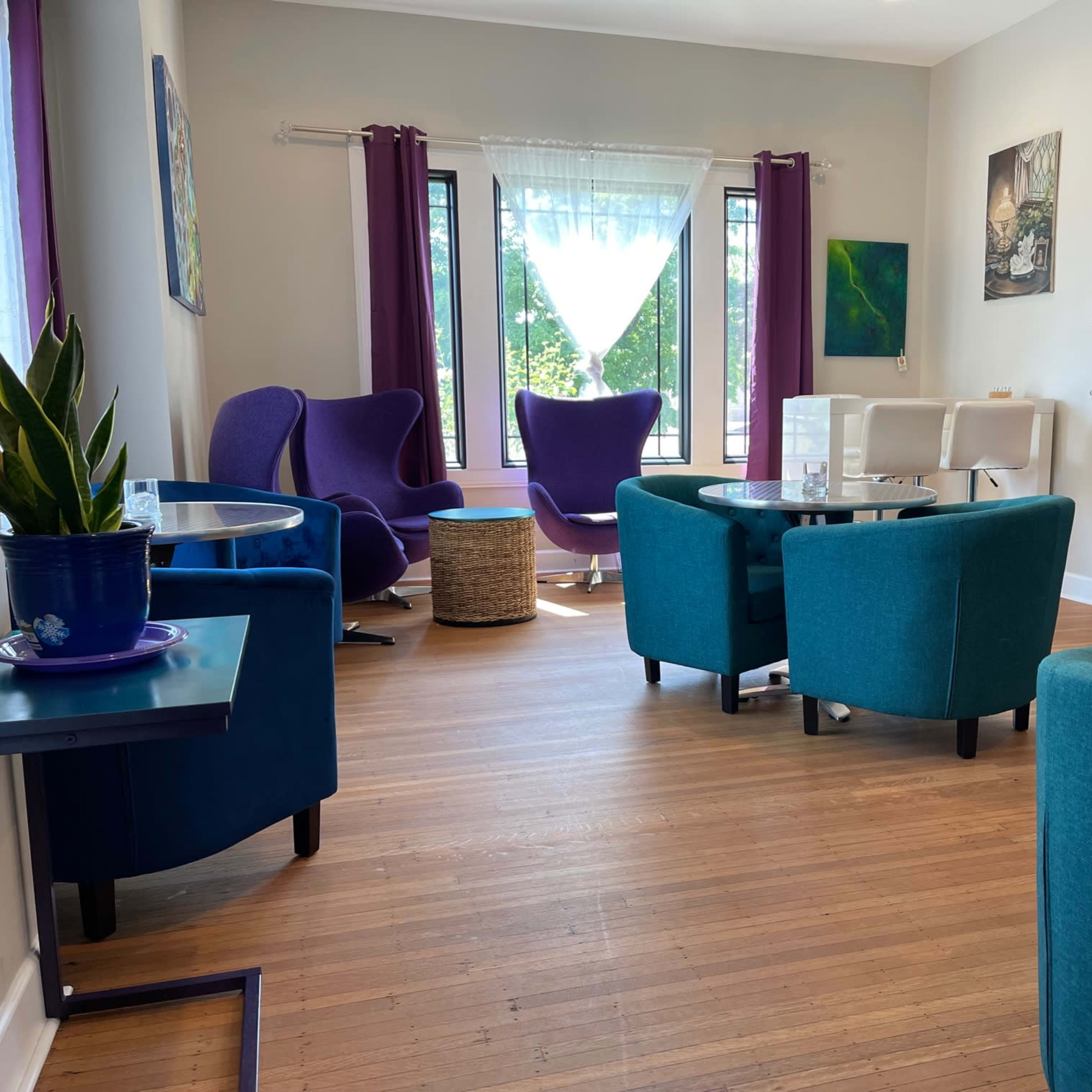 A bright room with multiple seating areas, featuring blue and purple chairs, a glass table, and large windows with light curtains.