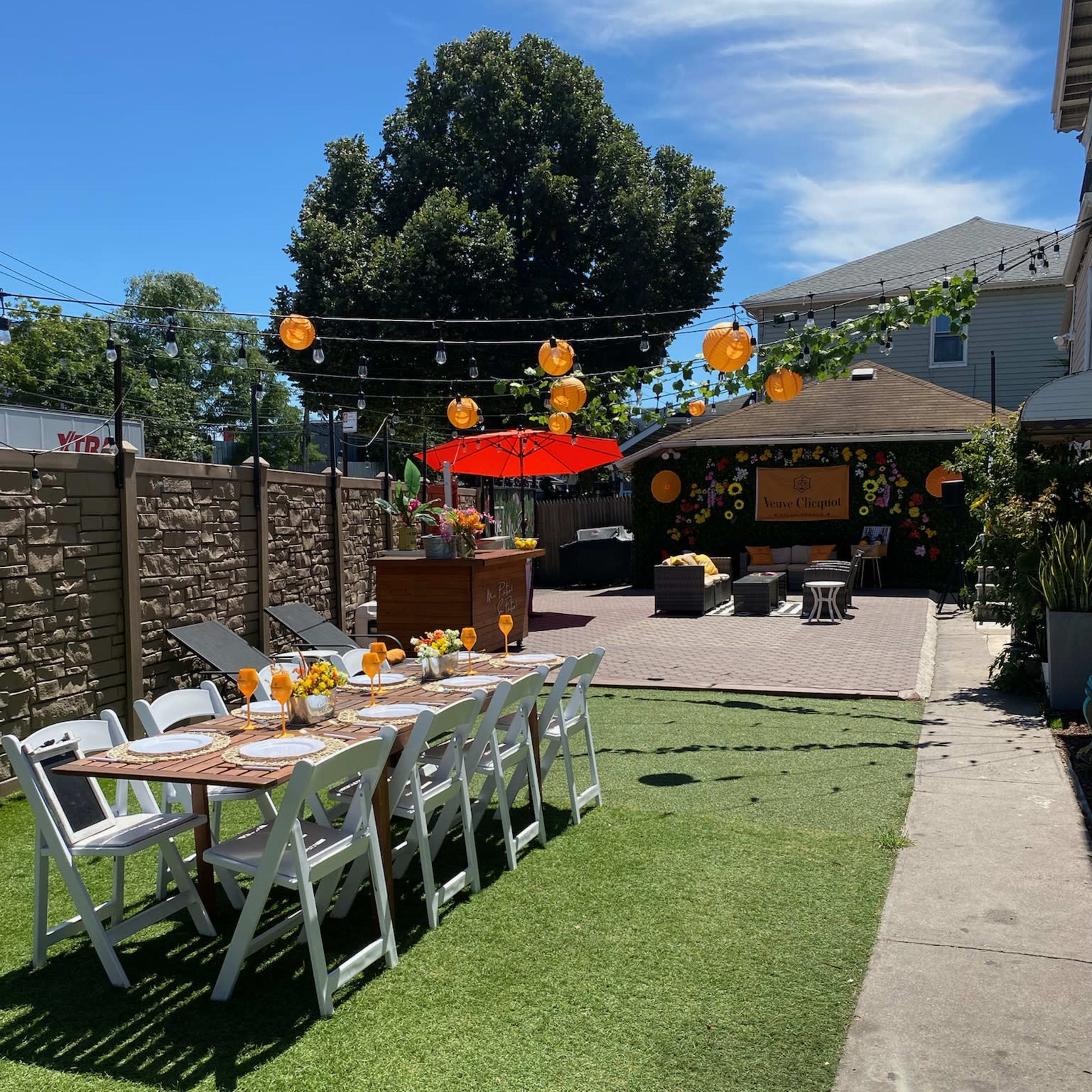 A decorated outdoor space features a long dining table set with flowers, surrounded by lush greenery and string lights.