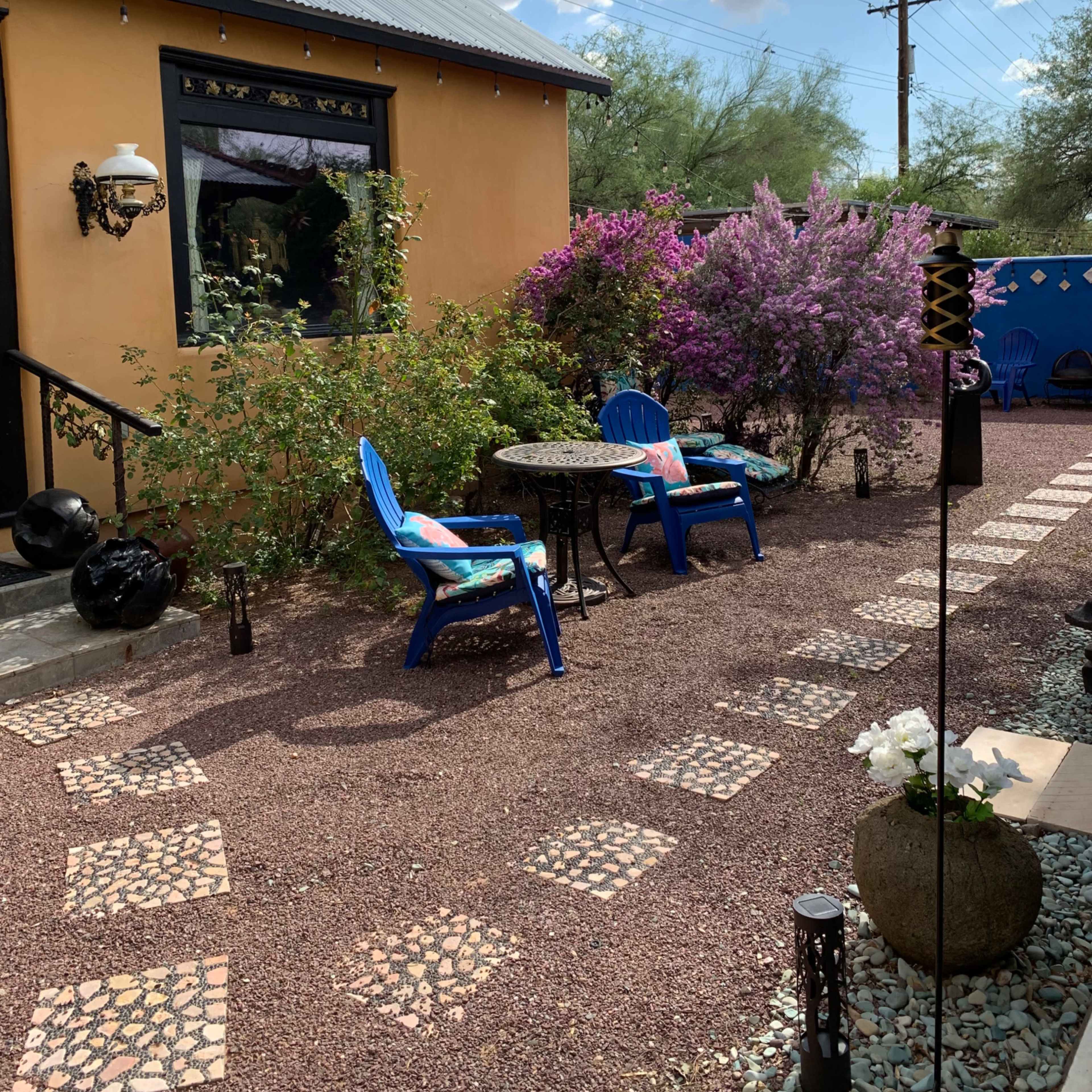 A colorful courtyard with a gravel pathway, two blue chairs at a small table, and vibrant flowering plants surrounding a building with a prominent window.