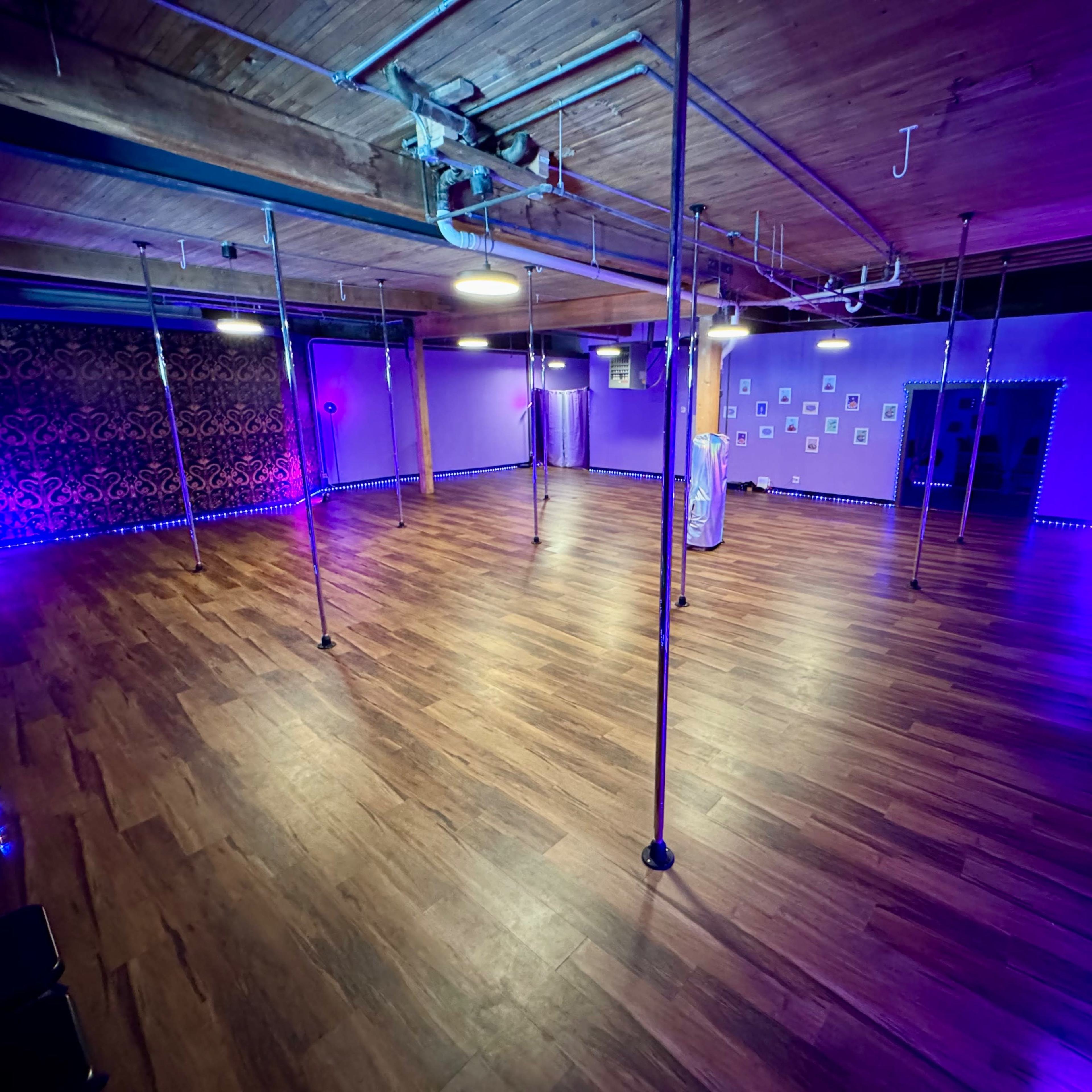 An empty dance studio with wooden flooring, multiple polished poles, and purple ambient lighting.
