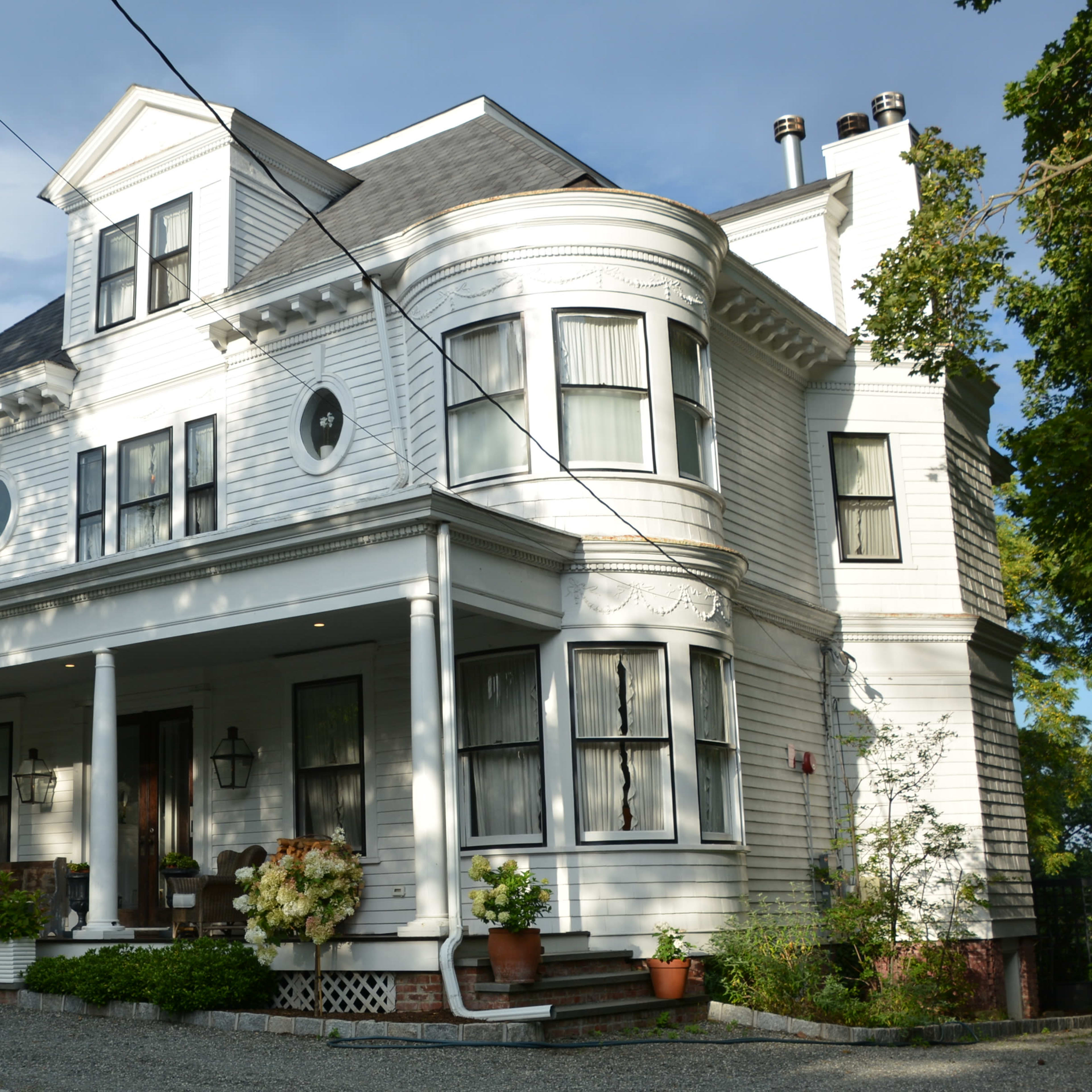 A large, two-story white house with a curved bay window and a front porch adorned with potted plants.