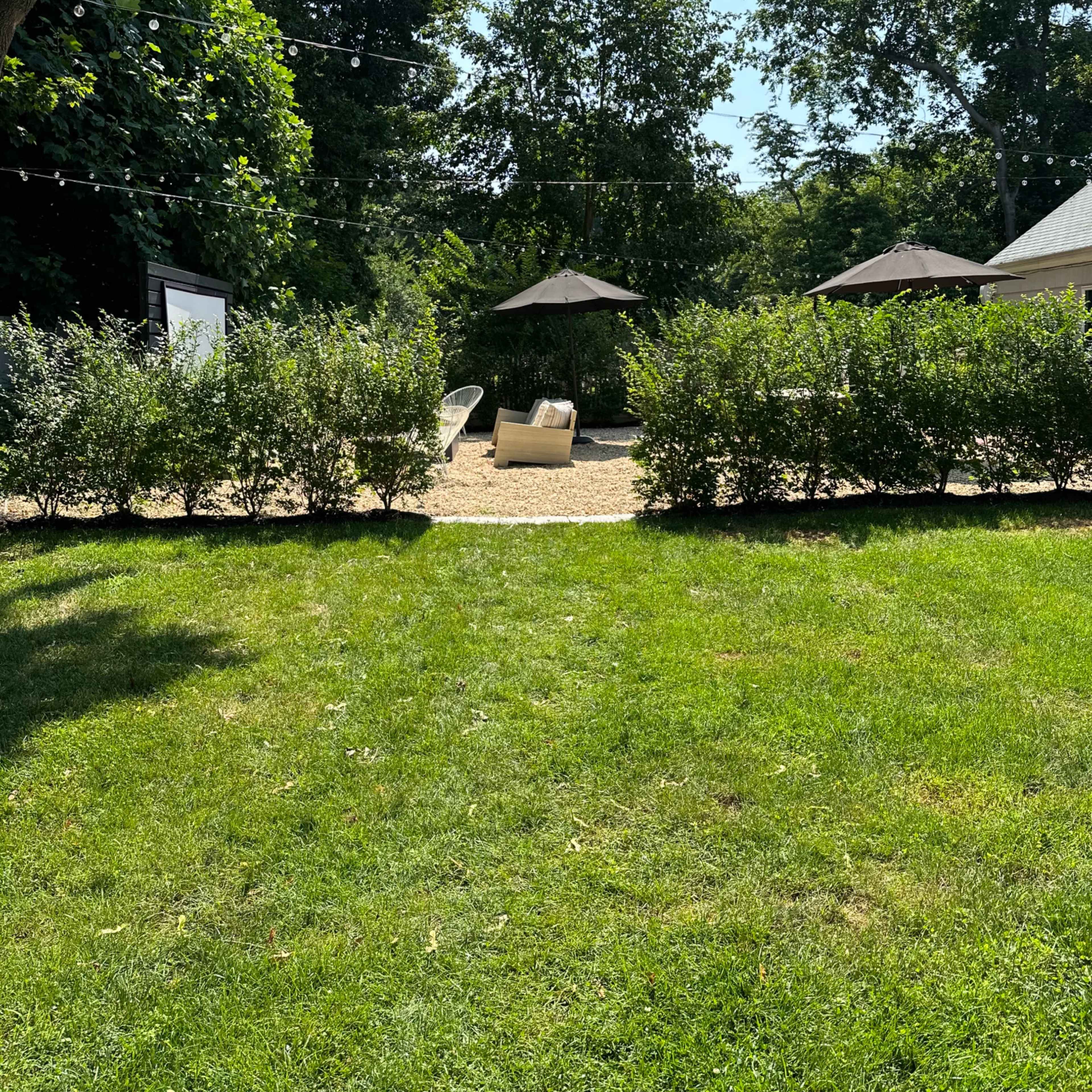 A landscaped area features a gravel path lined with shrubs, two lounge chairs under a canopy, and several trees in the background.