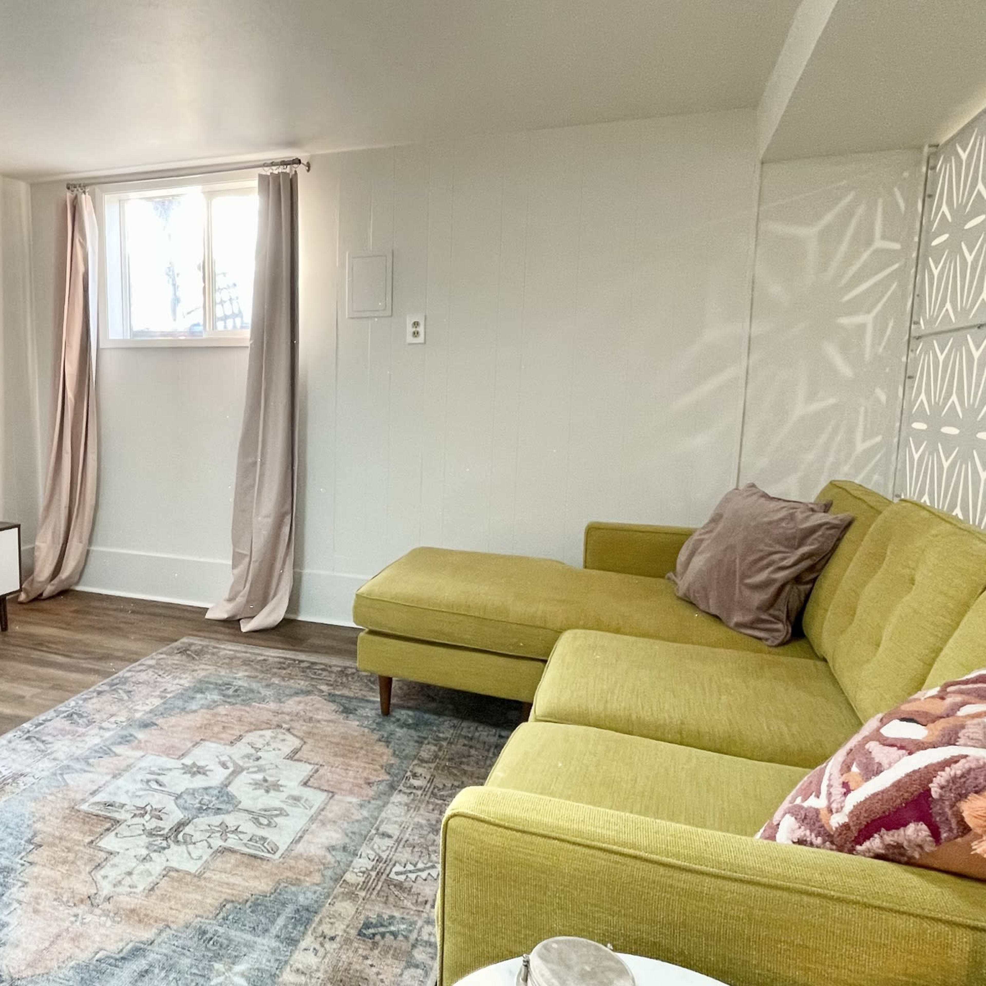 The scene shows a bright living room featuring a yellow sectional sofa, a wall-mounted television, and a patterned area rug.