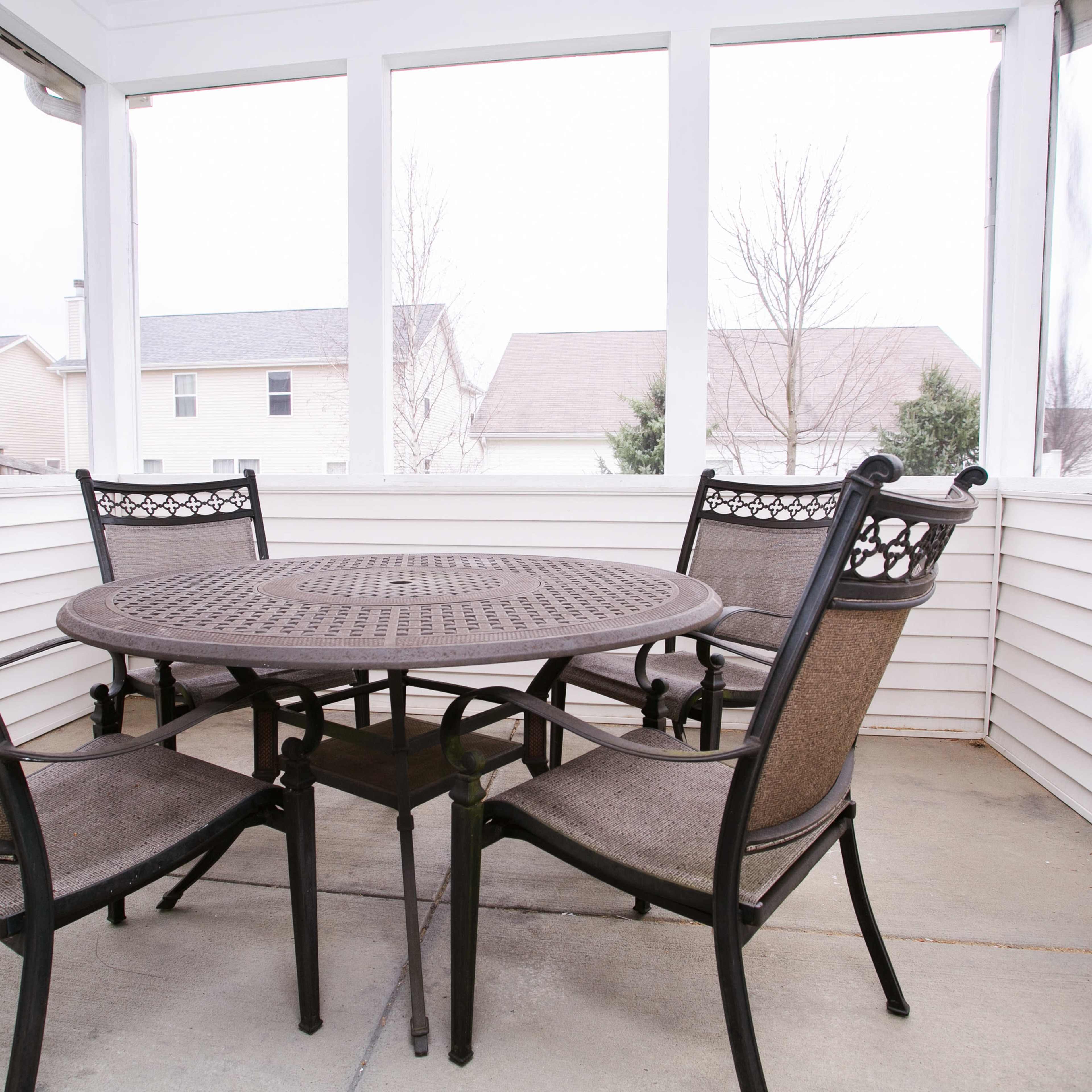 A round metal table with four chairs is set up in a screened-in porch surrounded by white walls.