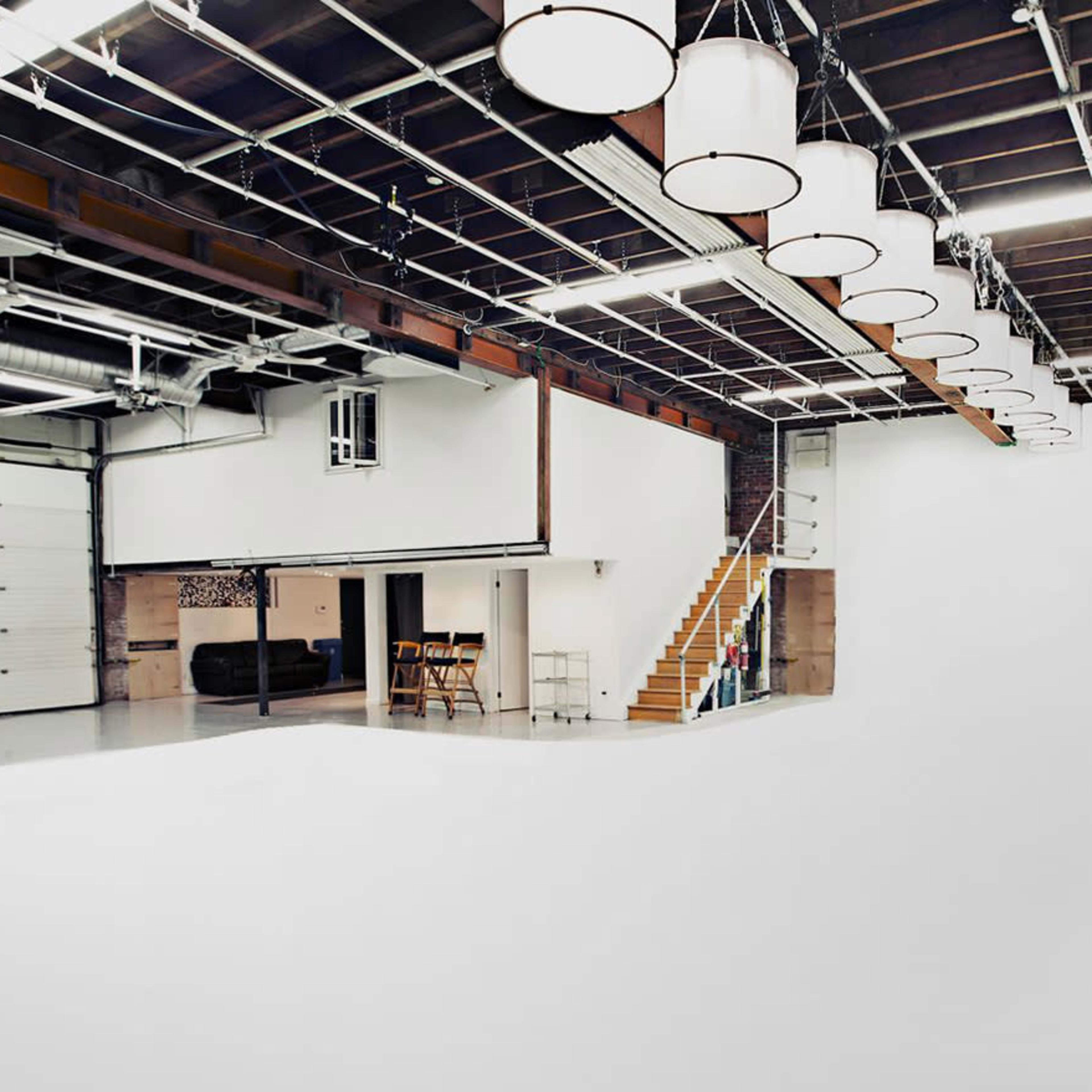 A spacious studio with a white cyclorama wall, overhead lighting, and a staircase leading to an upper level.