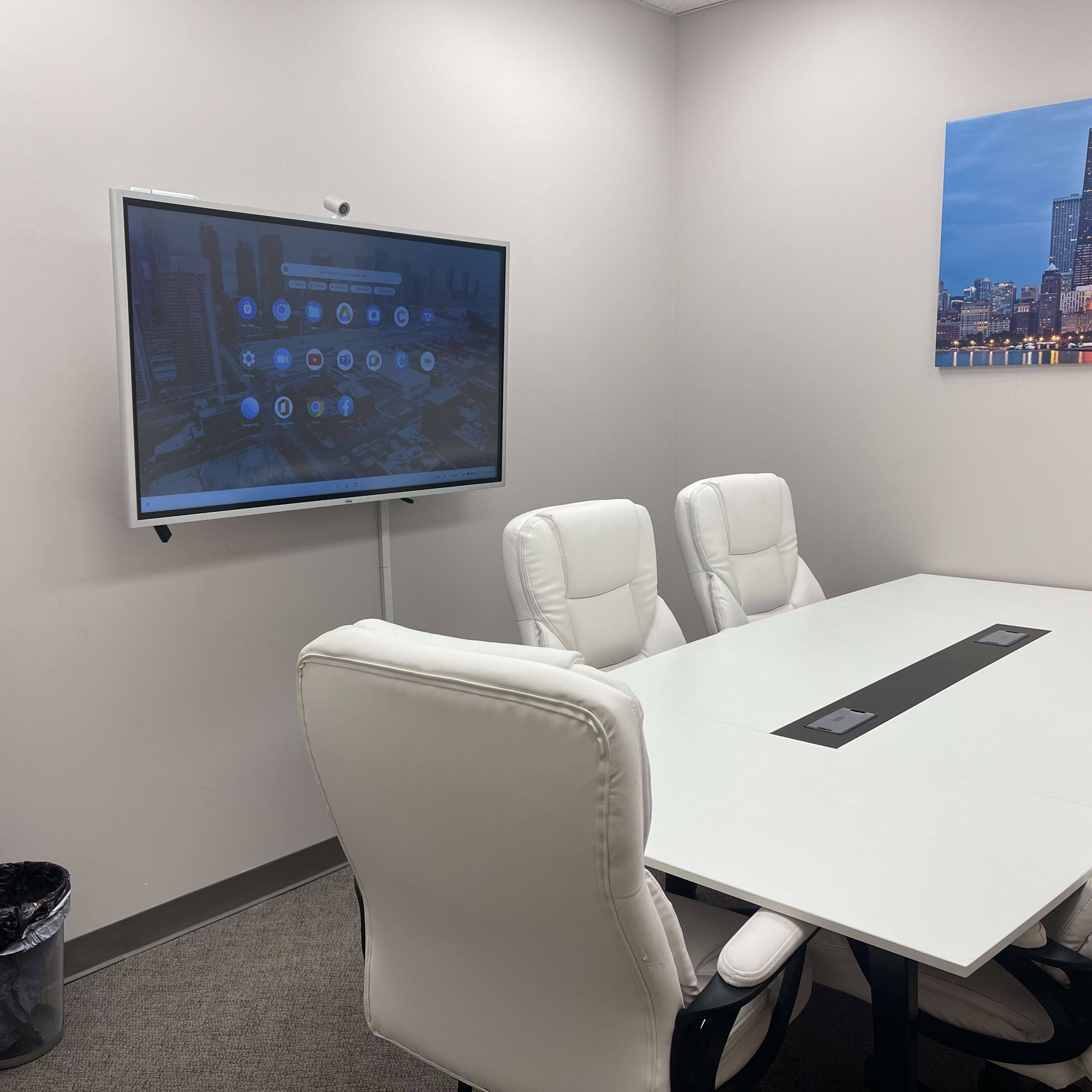 A modern conference room with a large screen displaying a digital interface, six white chairs around a rectangular table, and a small shelf with items in the corner.