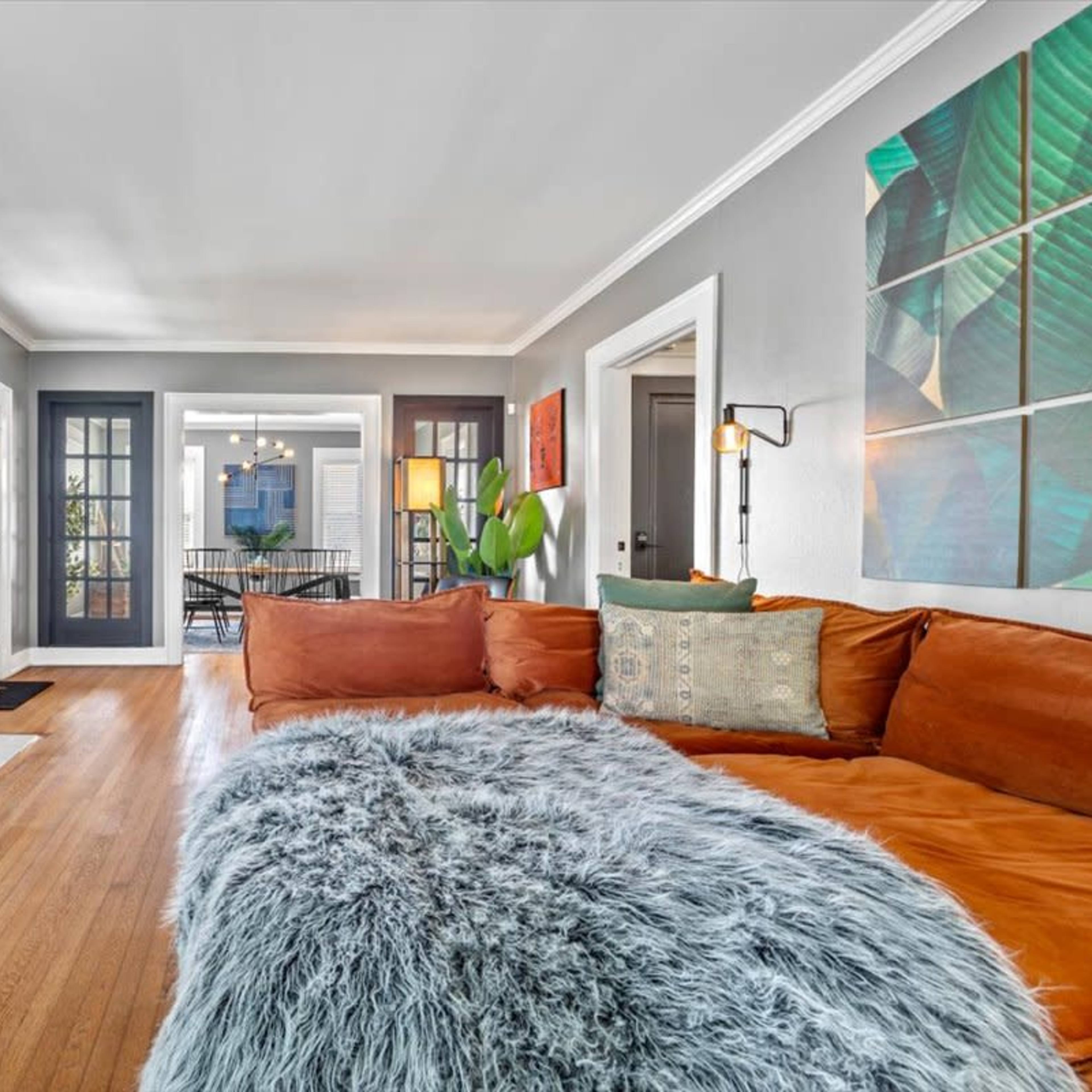 A cozy living room features a large orange sofa with a fluffy gray blanket, a wall-mounted TV, and doors leading to a dining area.