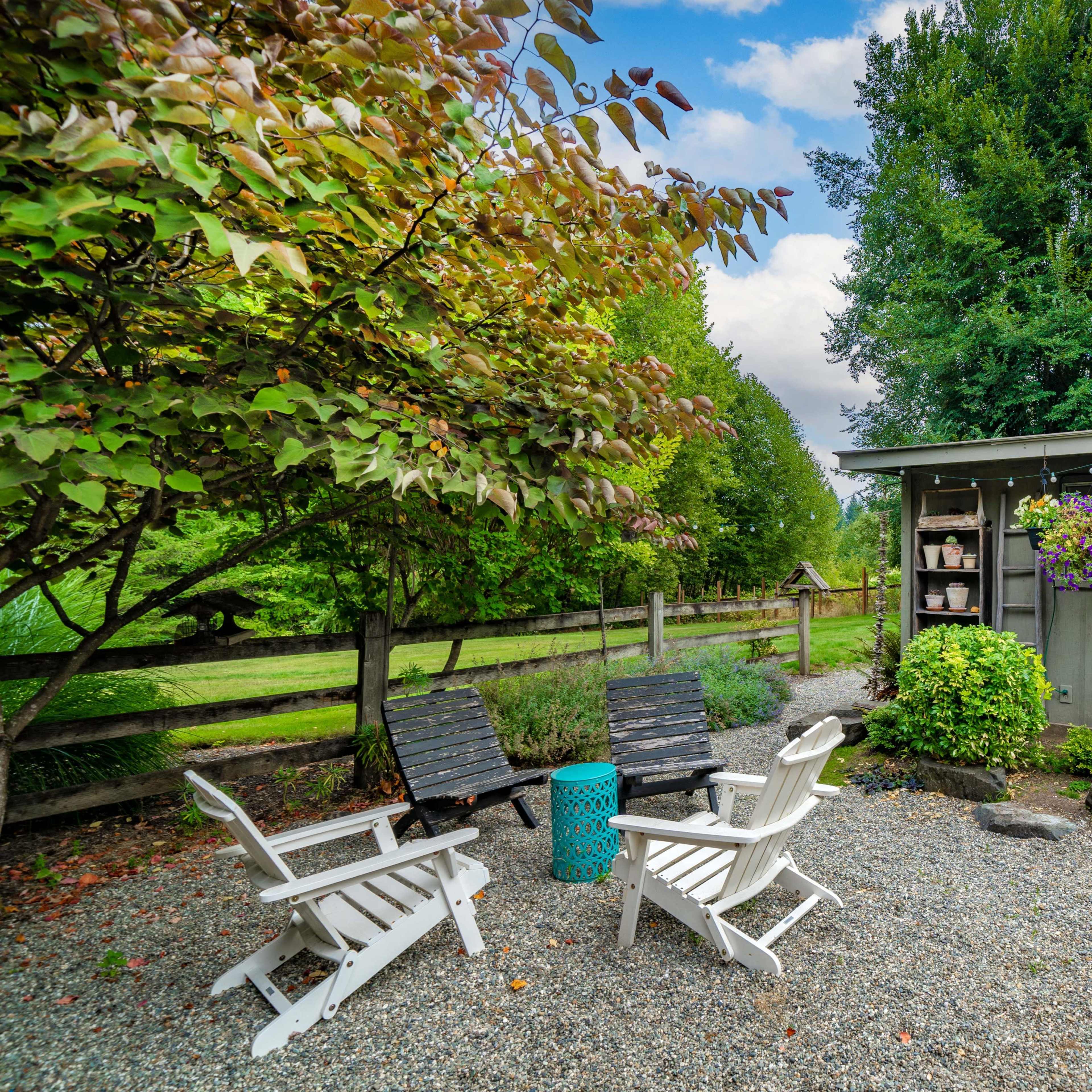 A gravel patio features four chairs arranged around a small table near a storage shed surrounded by greenery and flower pots.