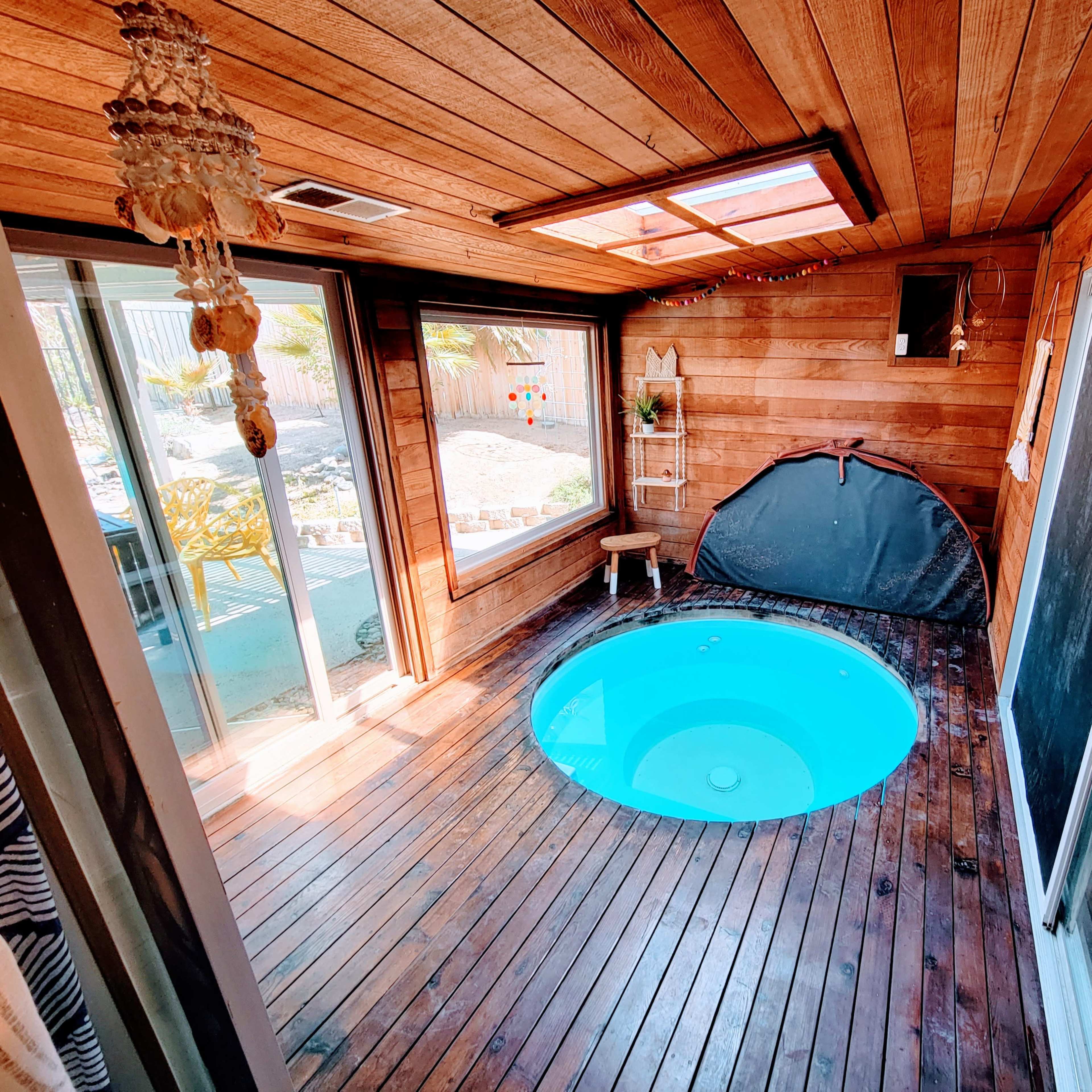 A wooden room with a circular hot tub, large windows, and a skylight, featuring wooden flooring and walls.