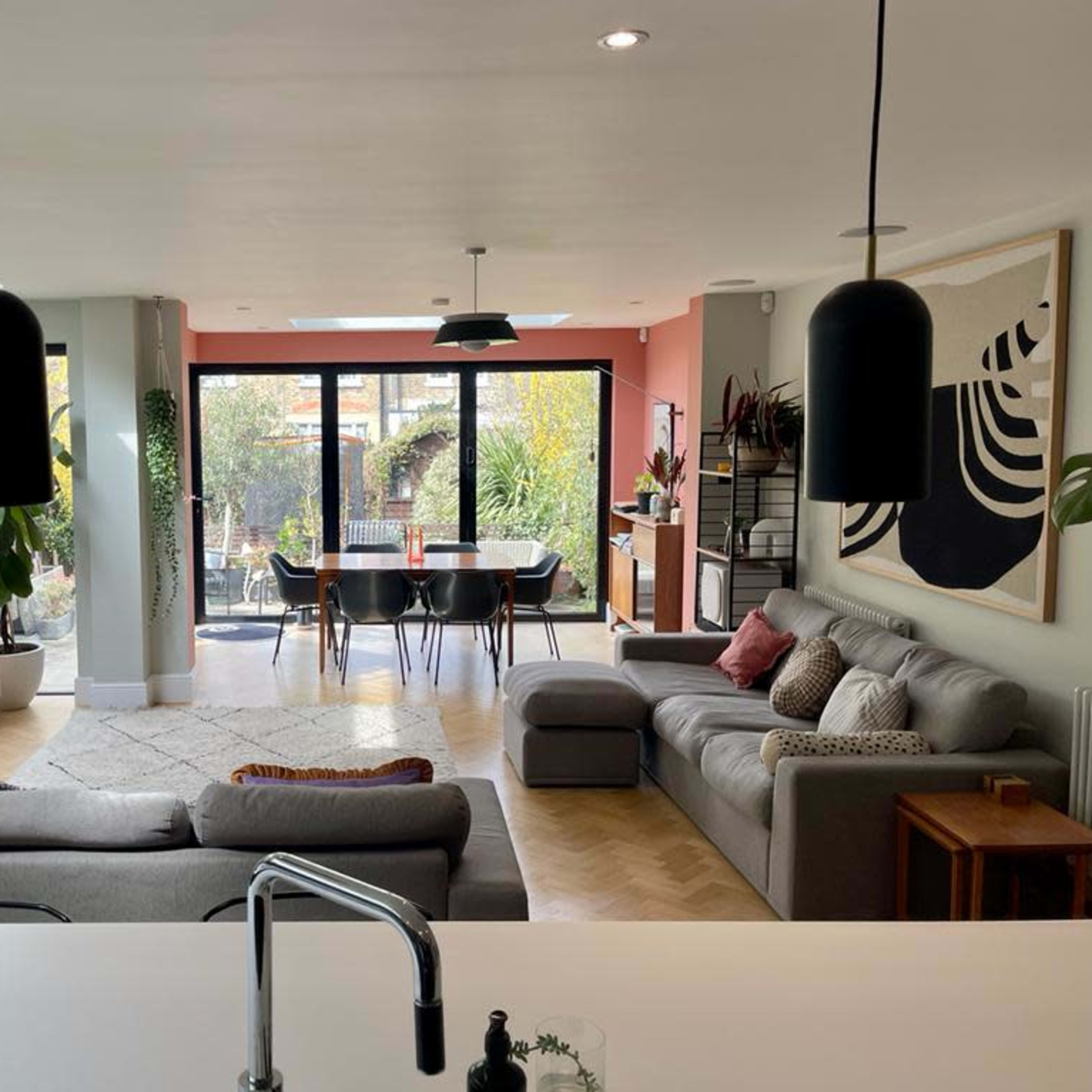 A modern living space features a sectional sofa, a dining area with a black table, and large sliding glass doors opening to a garden, surrounded by plants and contemporary decor.