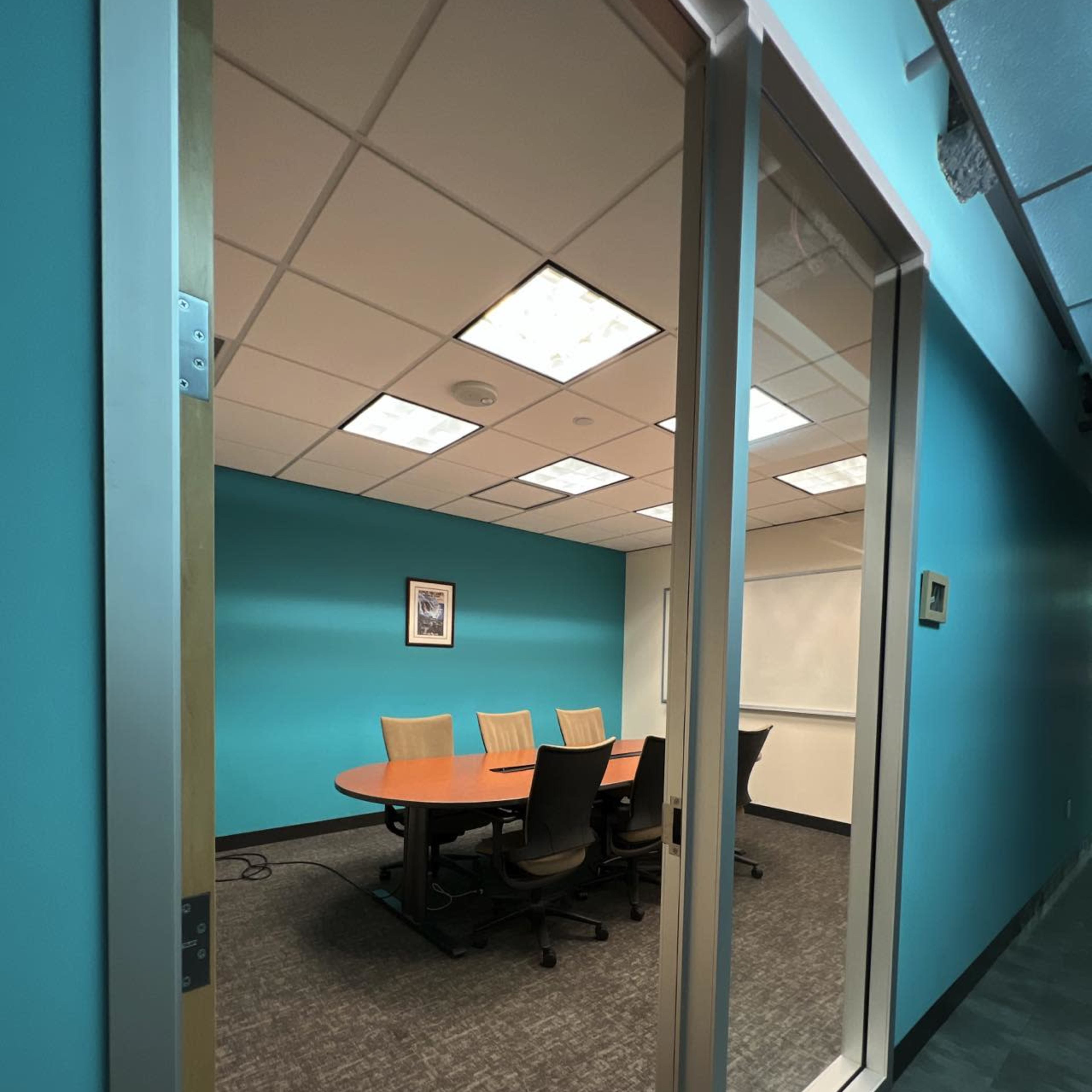 A conference room with a circular table and chairs is visible through a glass door, surrounded by turquoise walls and bright lighting.