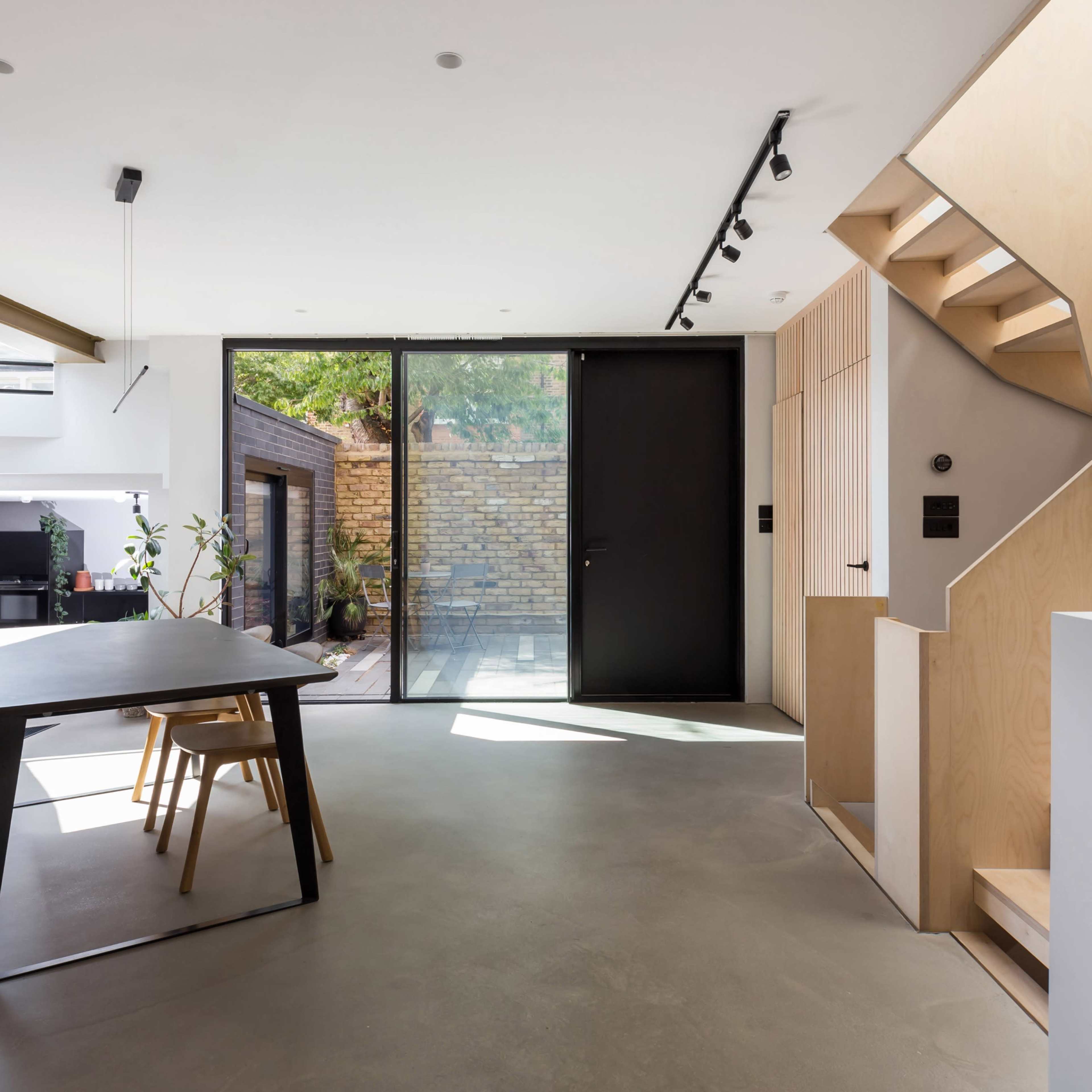 A modern interior space featuring a dining table with chairs, a staircase, a large glass door leading to a patio, and minimalistic decor.