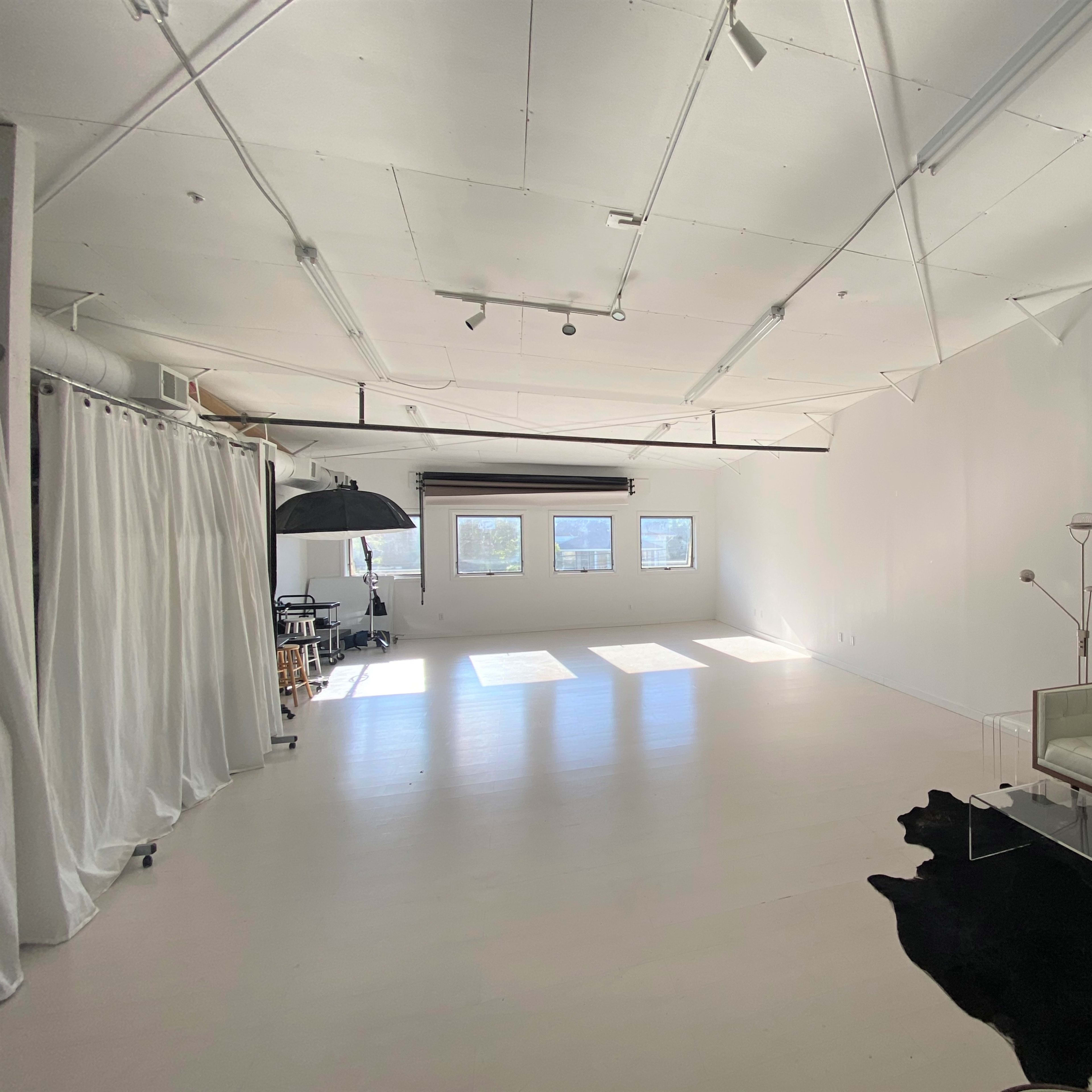 A spacious, minimalist studio with white walls, a large window, a black and white rug, a white sofa, and a modern floor lamp.