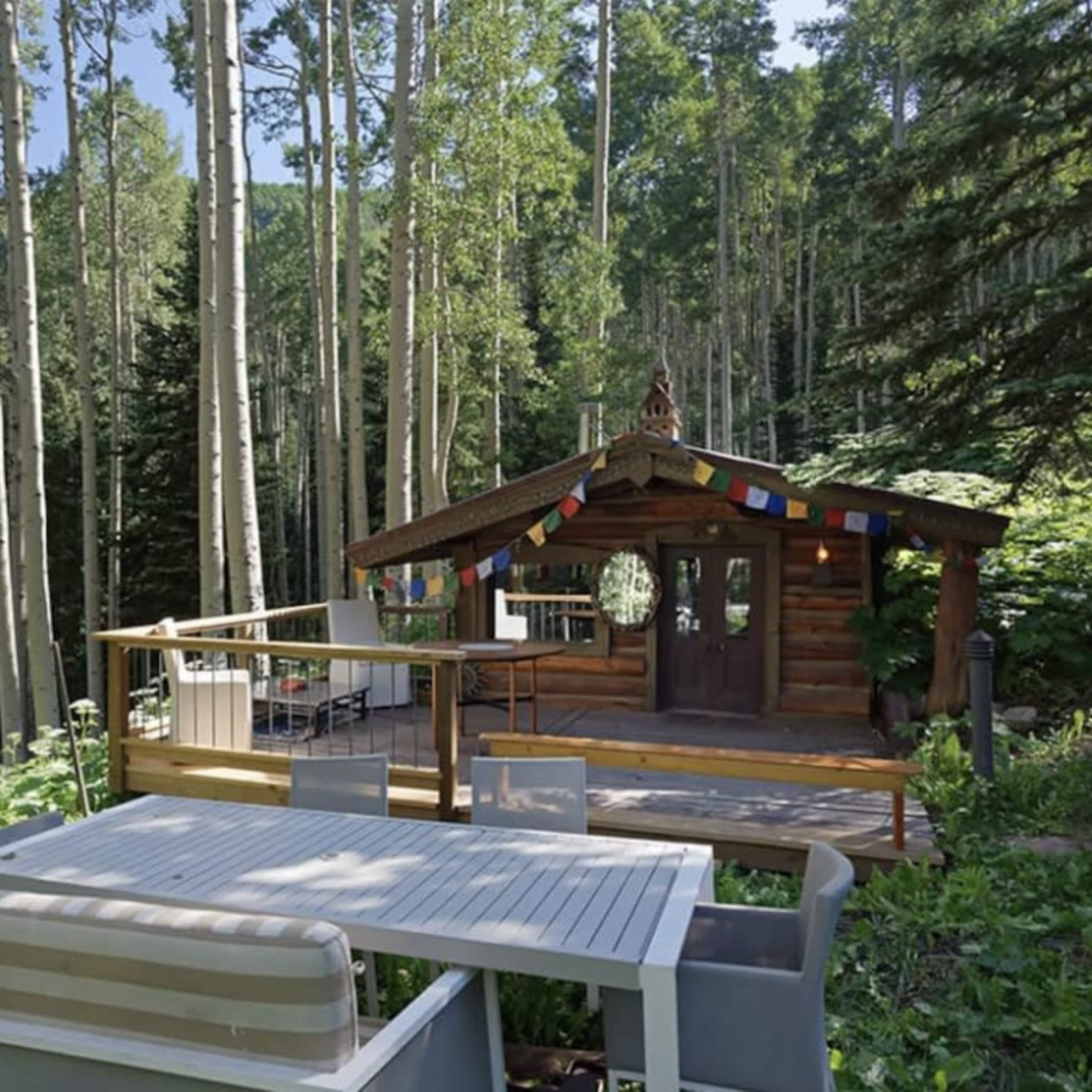 A wooden cabin surrounded by tall trees features an outdoor seating area with a white table and benches.