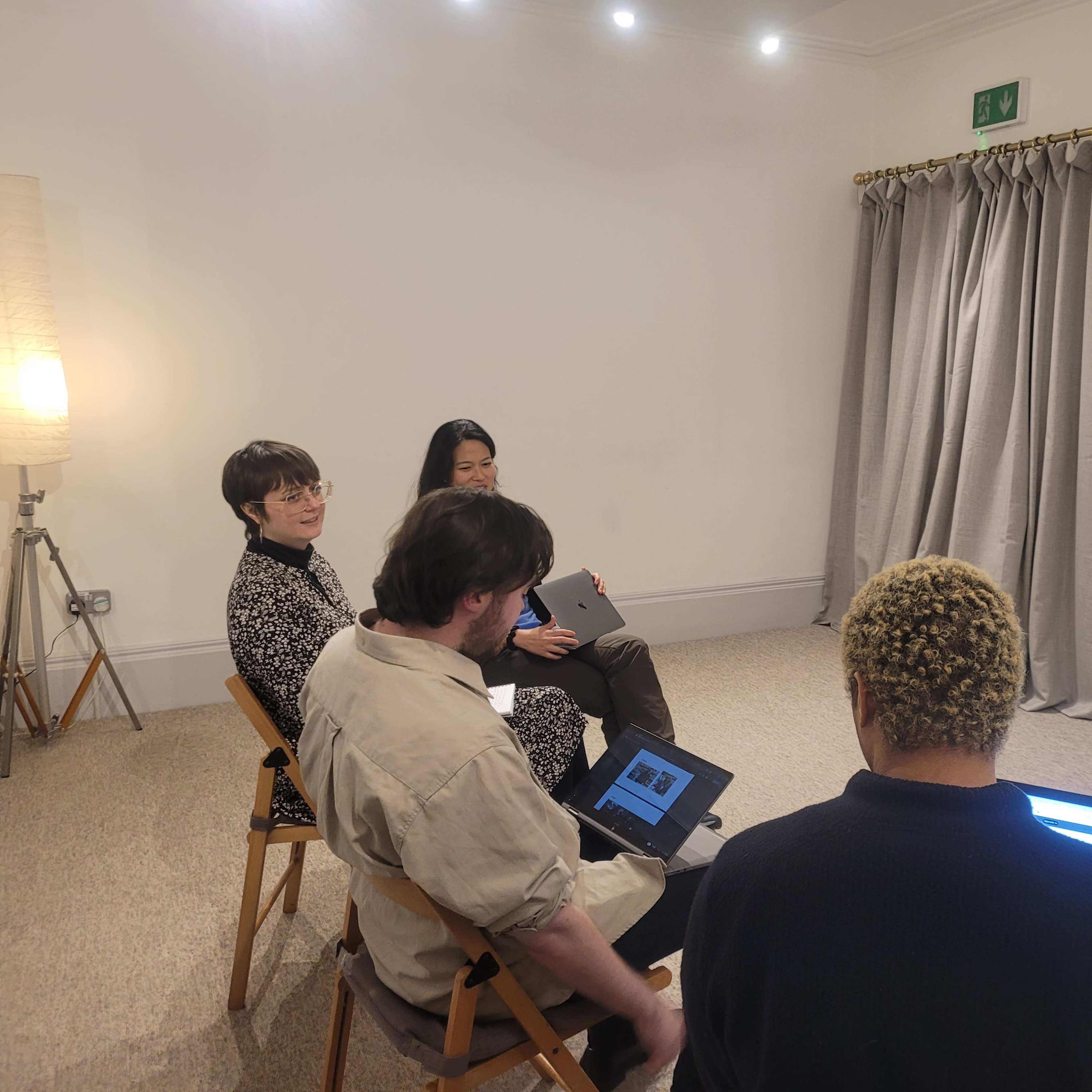 A group of four individuals sits in a circle in a well-lit room, facing a whiteboard with notes on it.