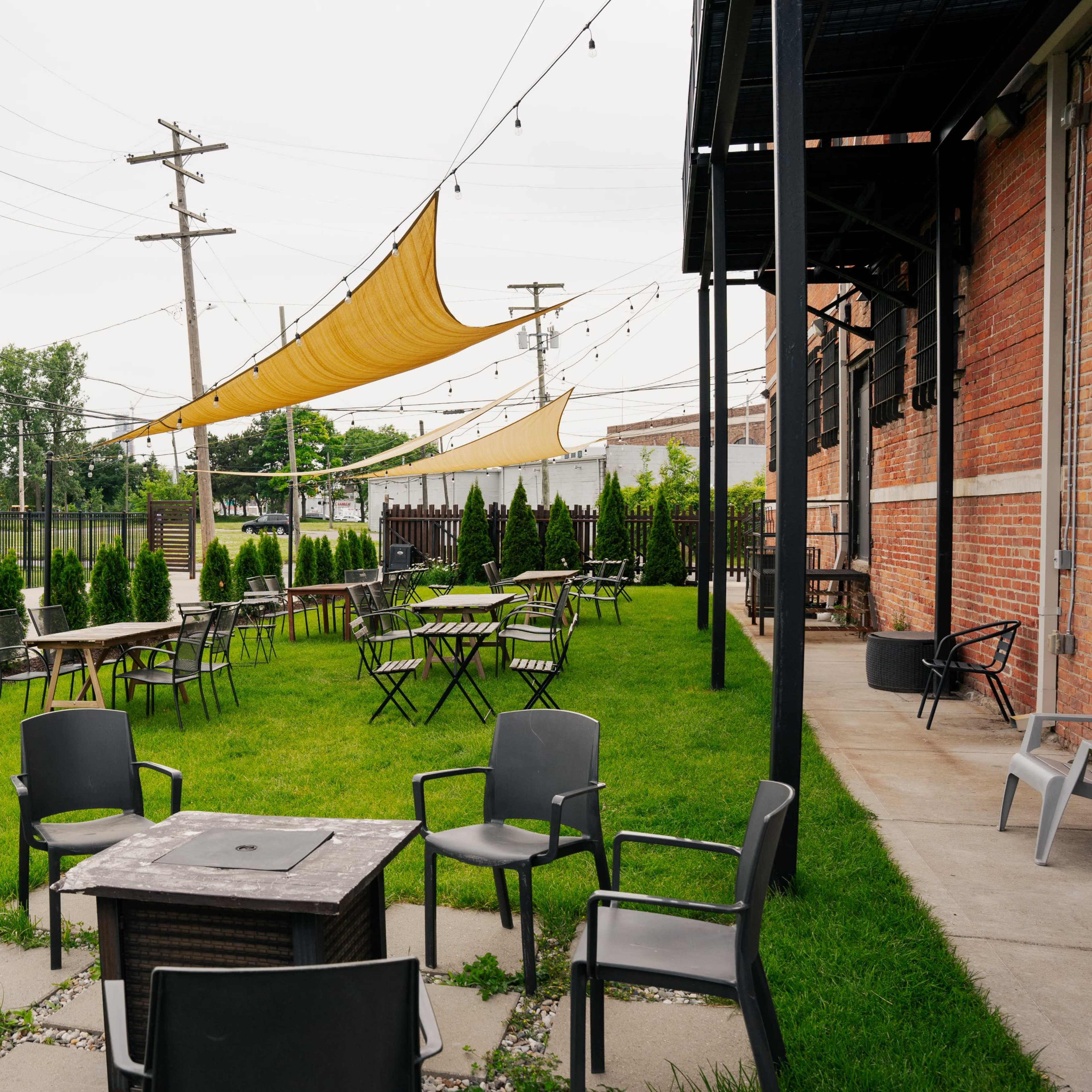 An outdoor seating area with empty metal chairs and tables arranged around a fire pit, surrounded by neatly trimmed grass and small trees, under yellow awnings.