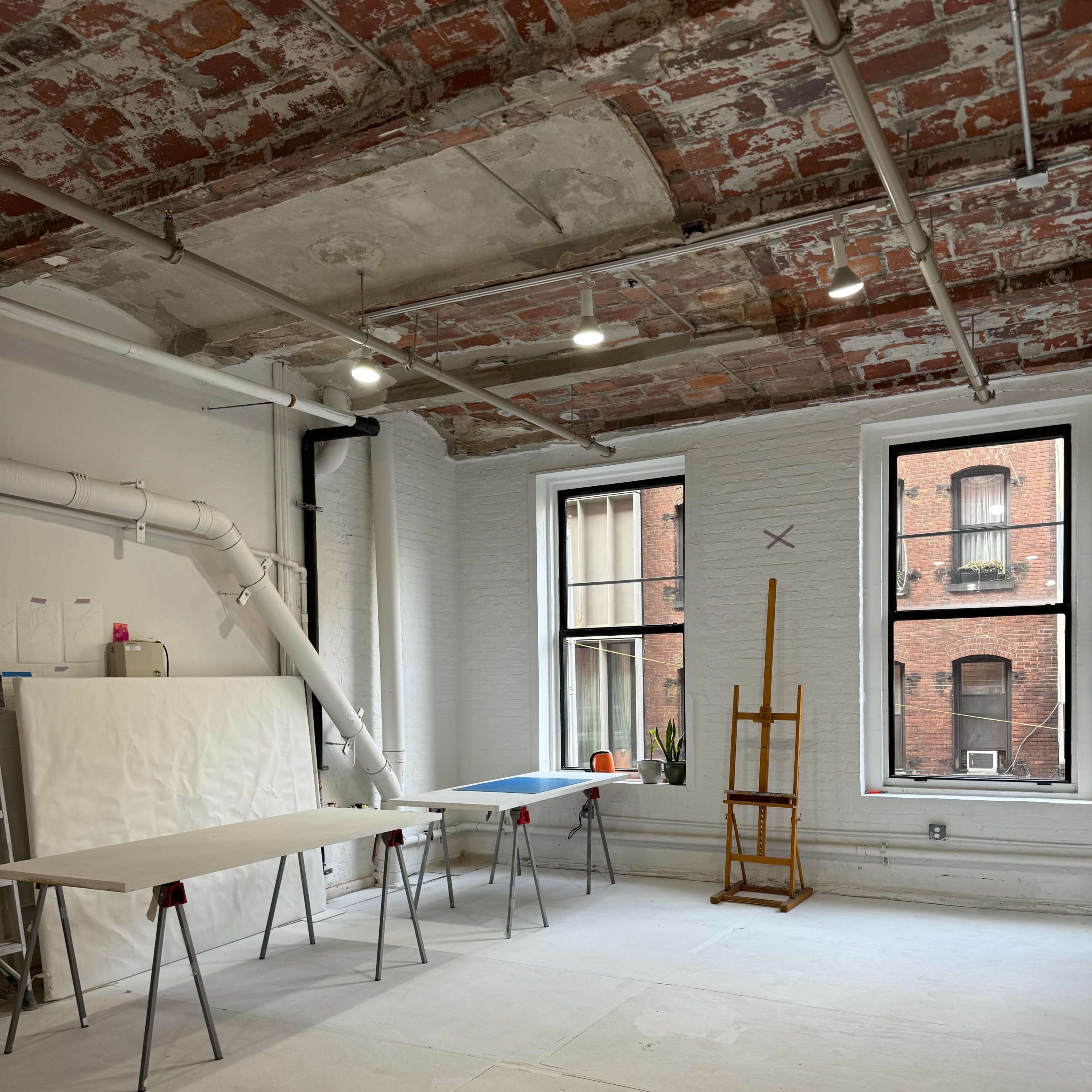 A spacious, well-lit studio features exposed brick walls, two large windows, a folding table, and an easel.