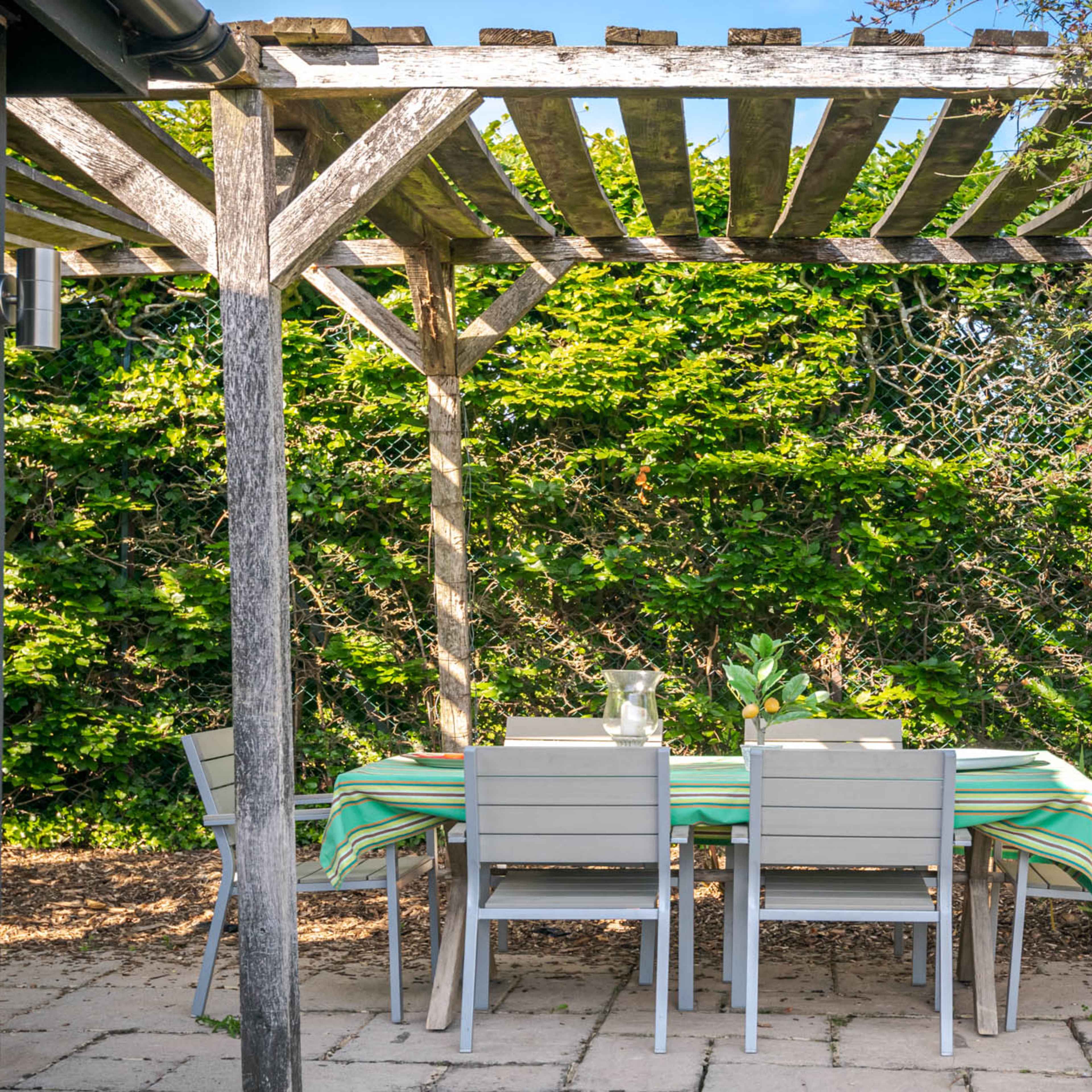 A patio area features a table and chairs set under a wooden pergola surrounded by greenery.