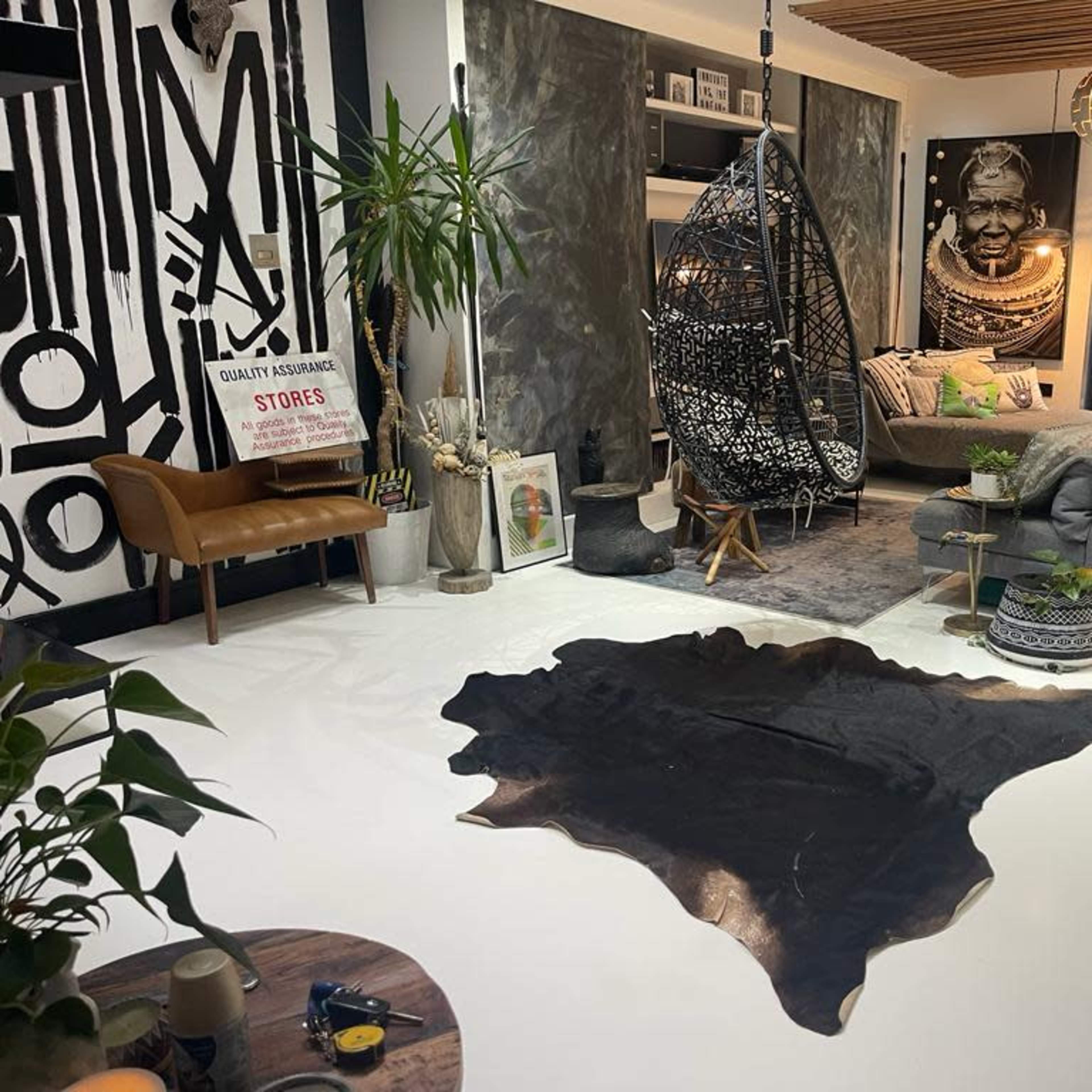 The room features a modern living space with a hanging wicker chair, a bed, a cowhide rug, and various indoor plants against a backdrop of decorative wall art.