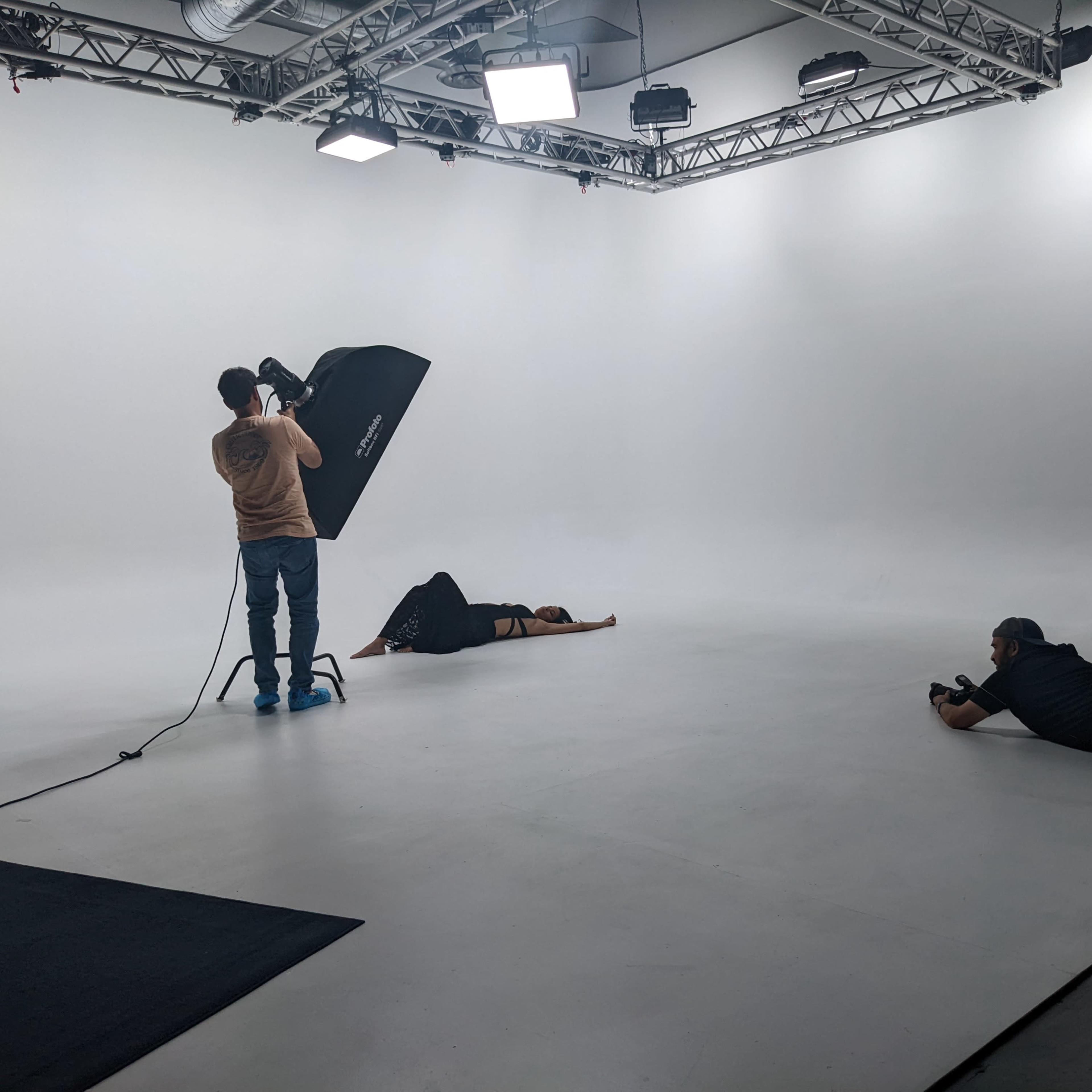 A photographer uses a softbox while a model lies on the ground in a white studio with two assistants positioned around them.