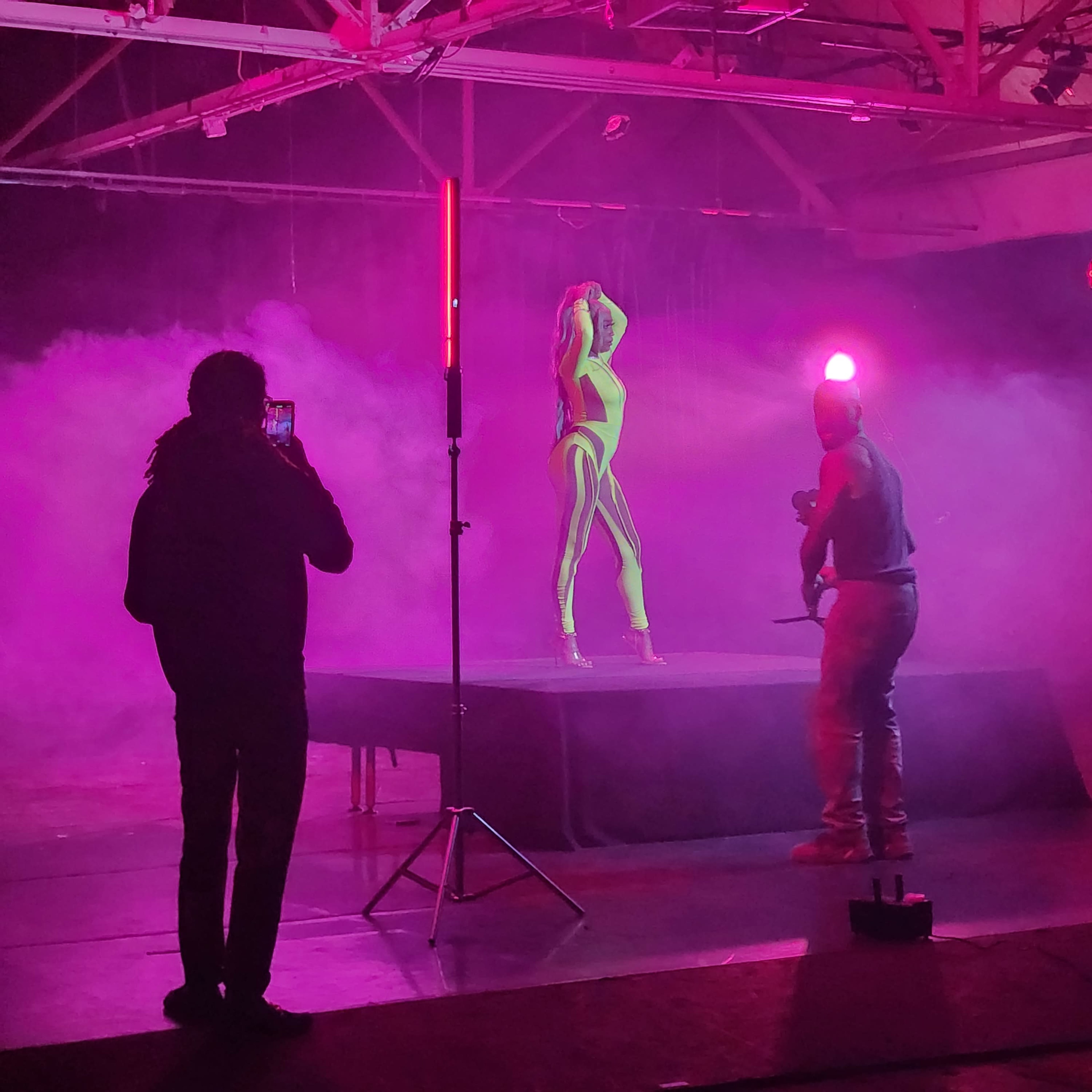A model poses on a platform while a photographer captures images in a studio filled with colored smoke and dramatic lighting.
