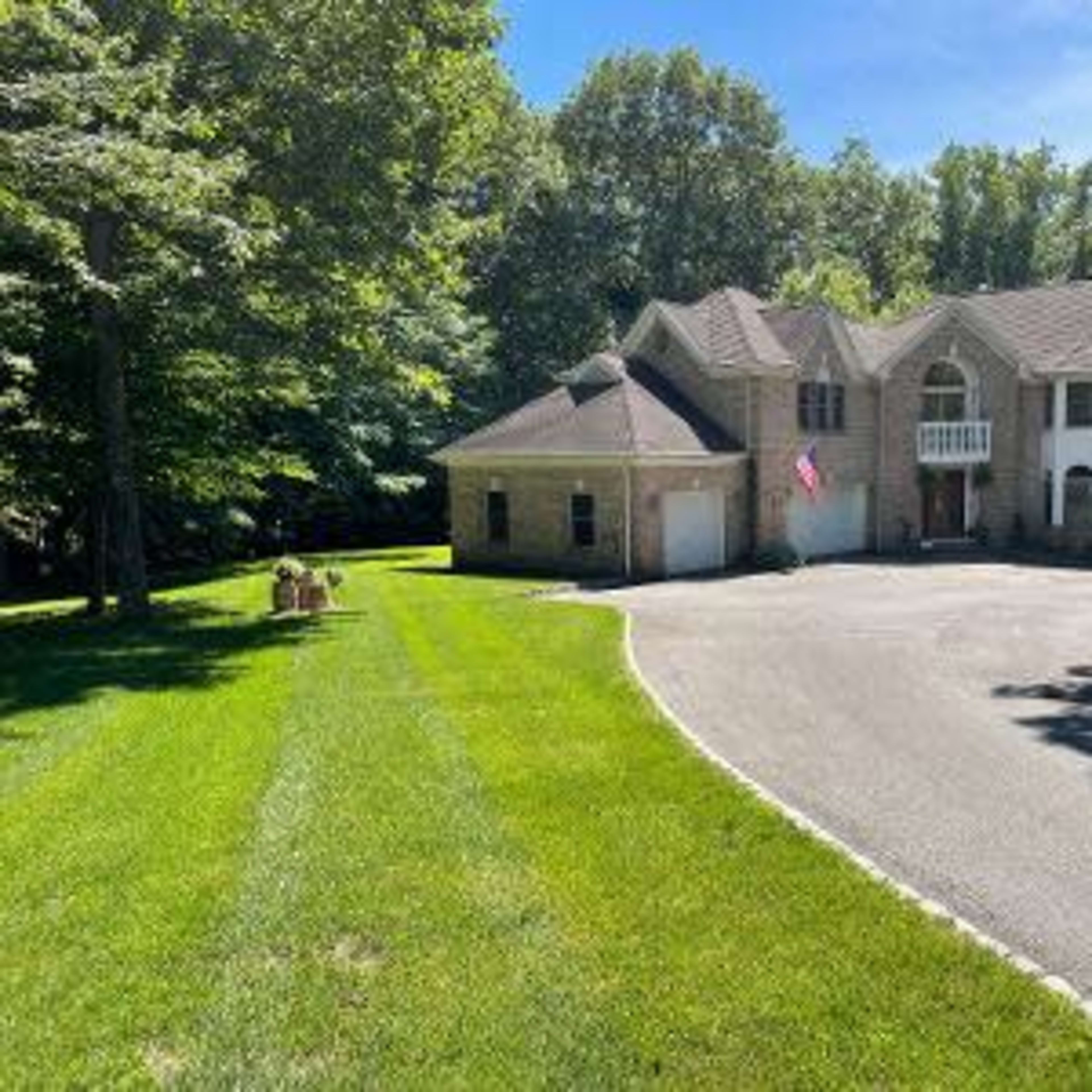 A large house with stone facade and a front yard is situated on a paved driveway surrounded by trees.