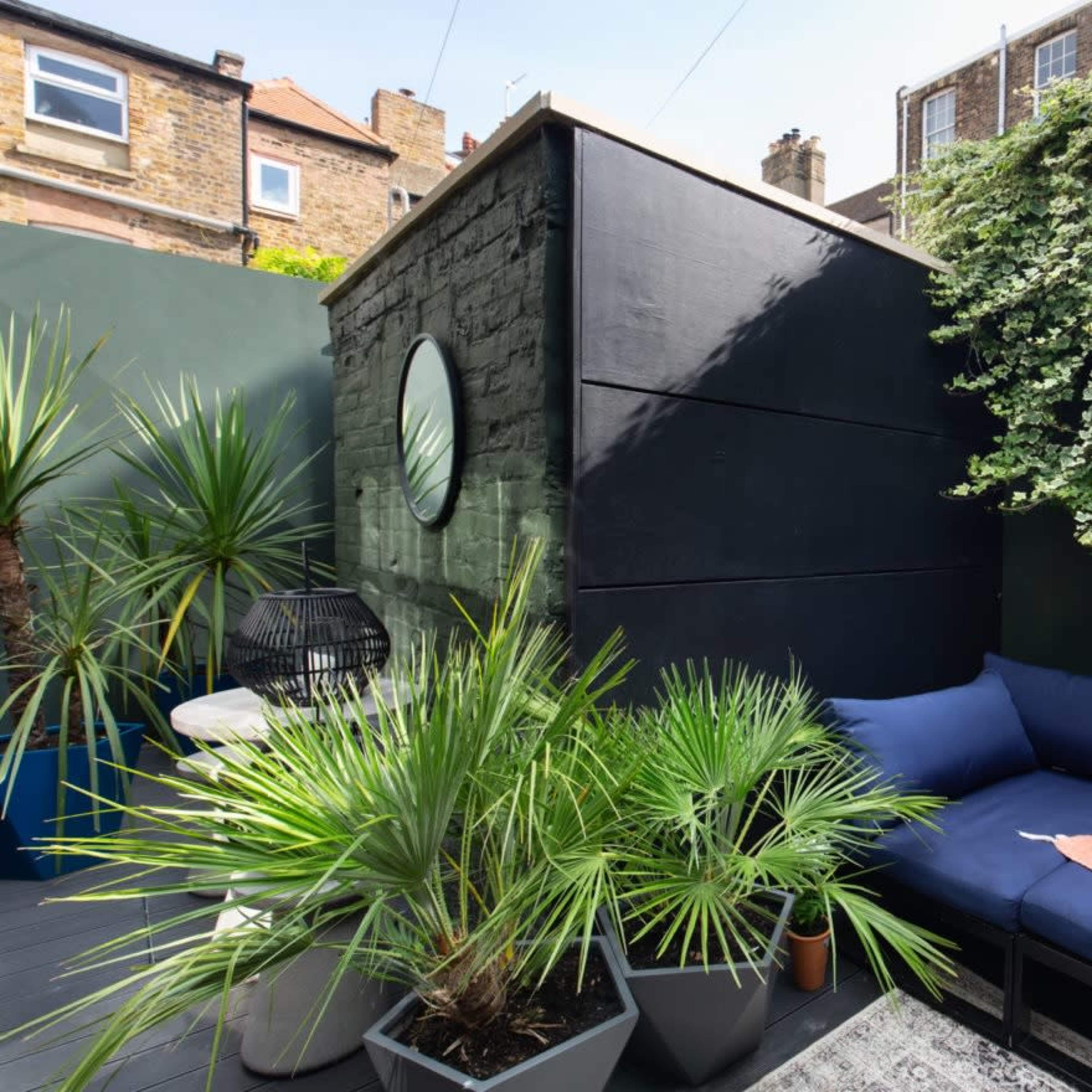 A small outdoor space with a black wall, a mirror, a seating area with blue cushions, and various potted plants.