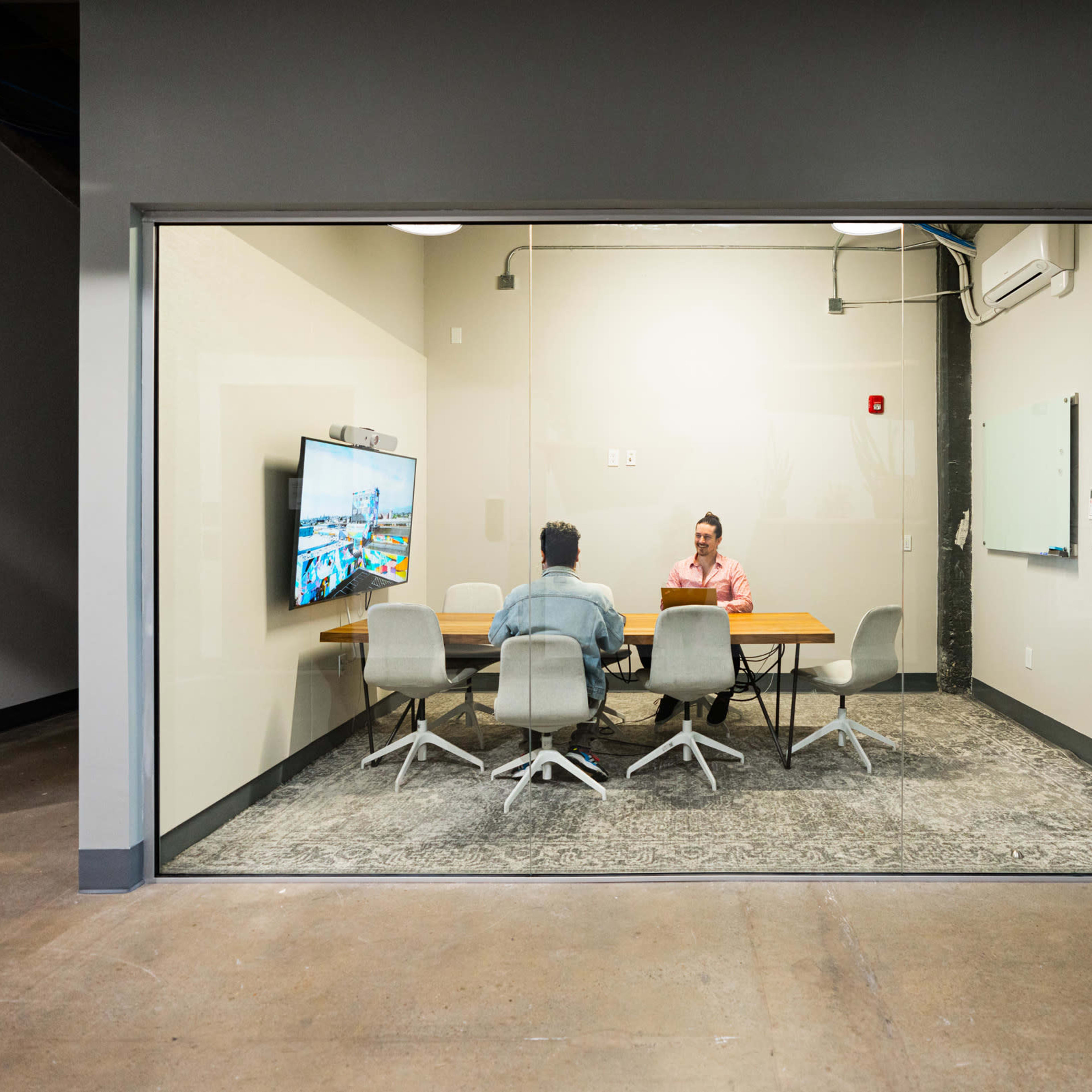 Two people are seated at a conference table in a glass-walled meeting room, with a large screen displaying a digital presentation.