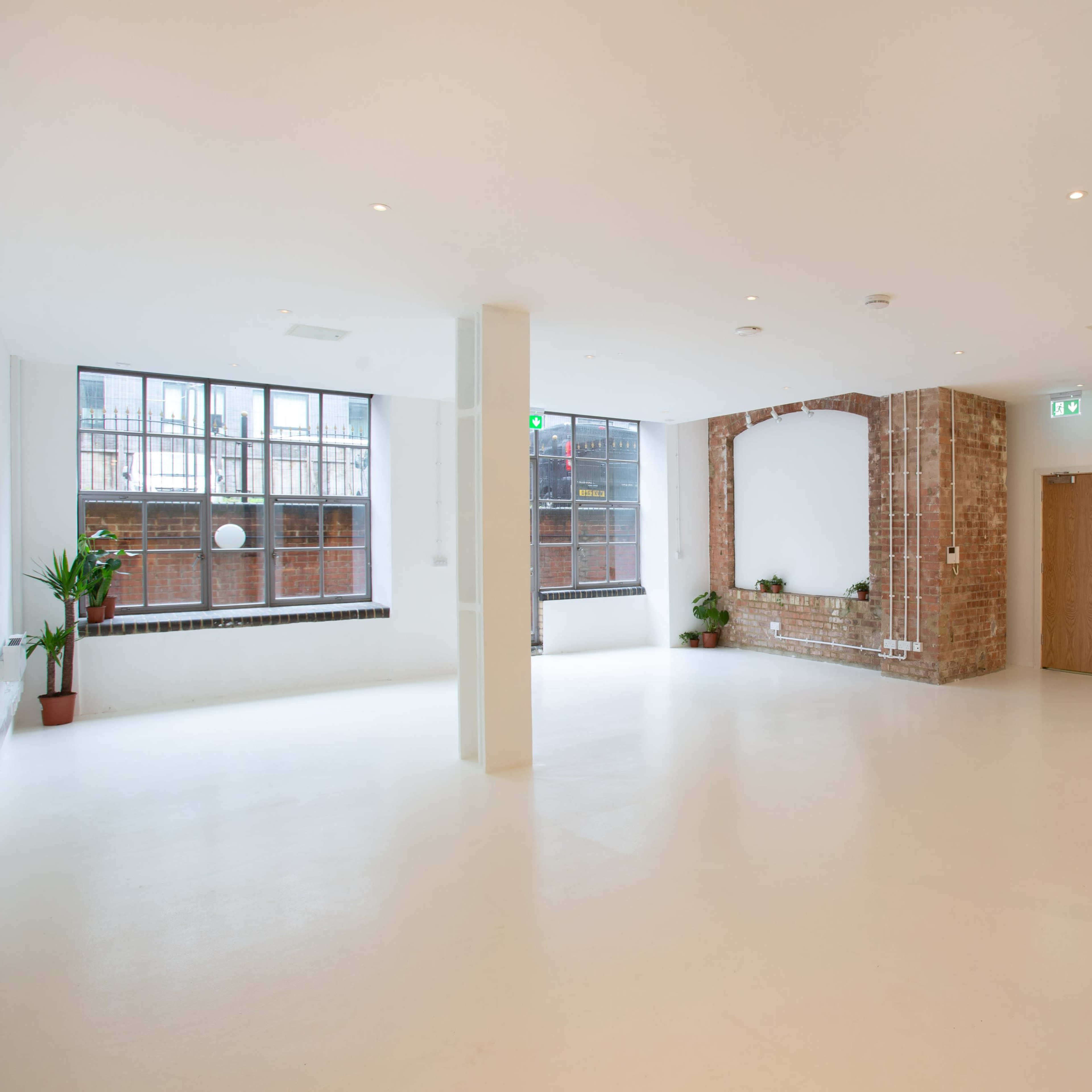 A spacious, empty room with white walls, a brick accent wall, large windows, and minimal furniture.