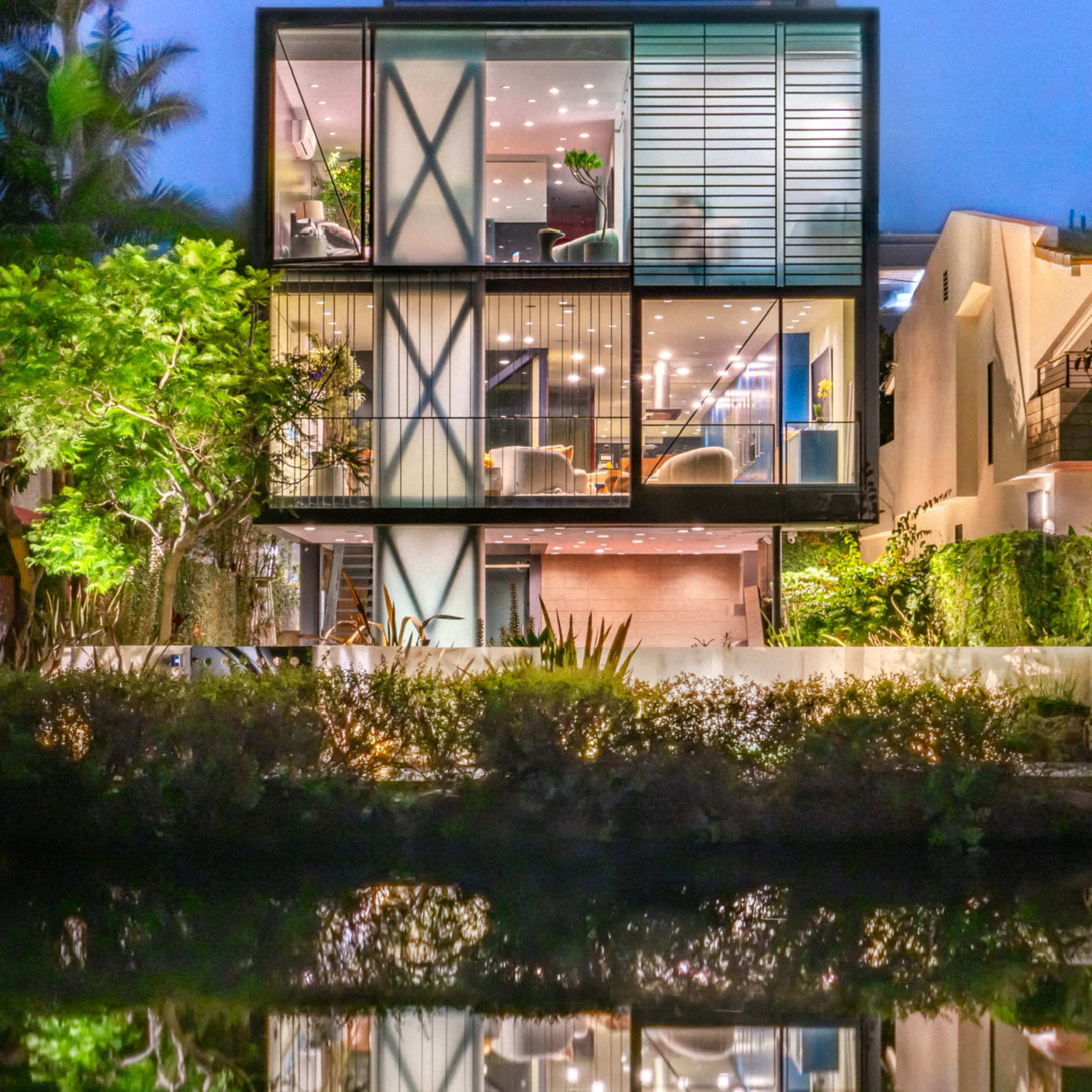 A modern two-story house with large glass windows and a reflective water feature in front showcases a sleek architectural design surrounded by greenery.