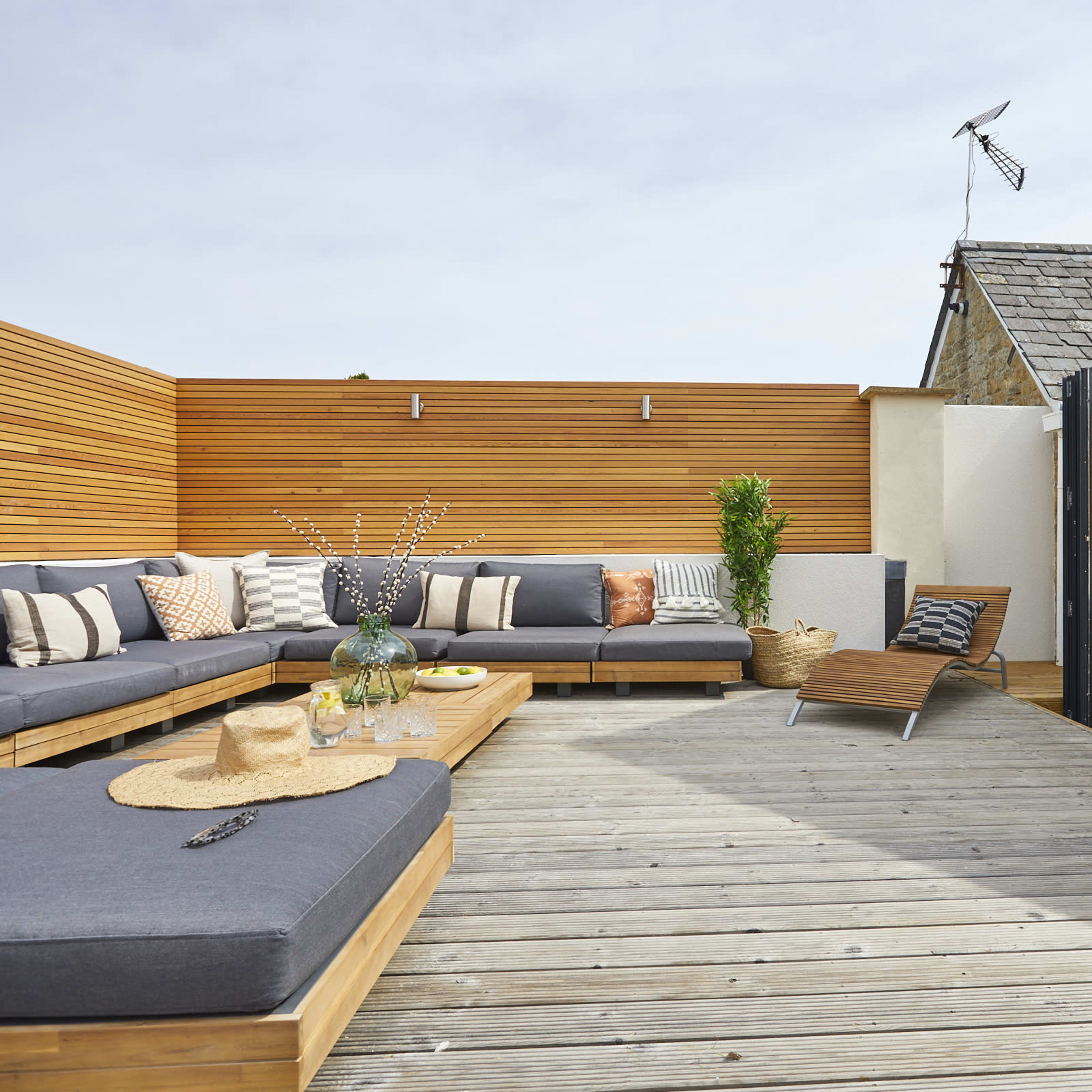 A modern outdoor lounge area features a wooden deck with sectional seating, a coffee table, and a sun lounger, surrounded by wooden privacy panels and potted plants.