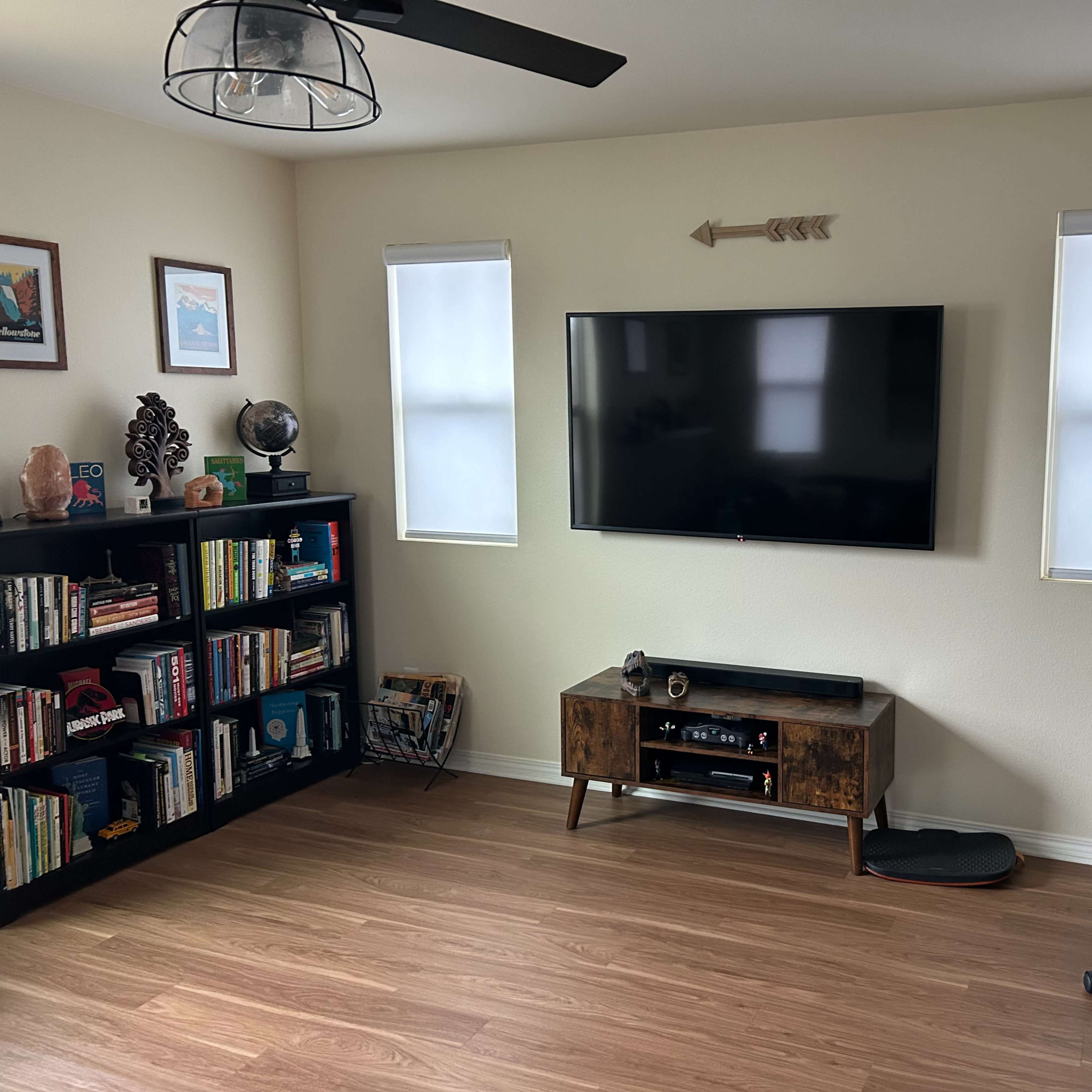 A living room features a large television mounted on the wall, flanked by windows, with a bookshelf and a desk with a chair in the corner.
