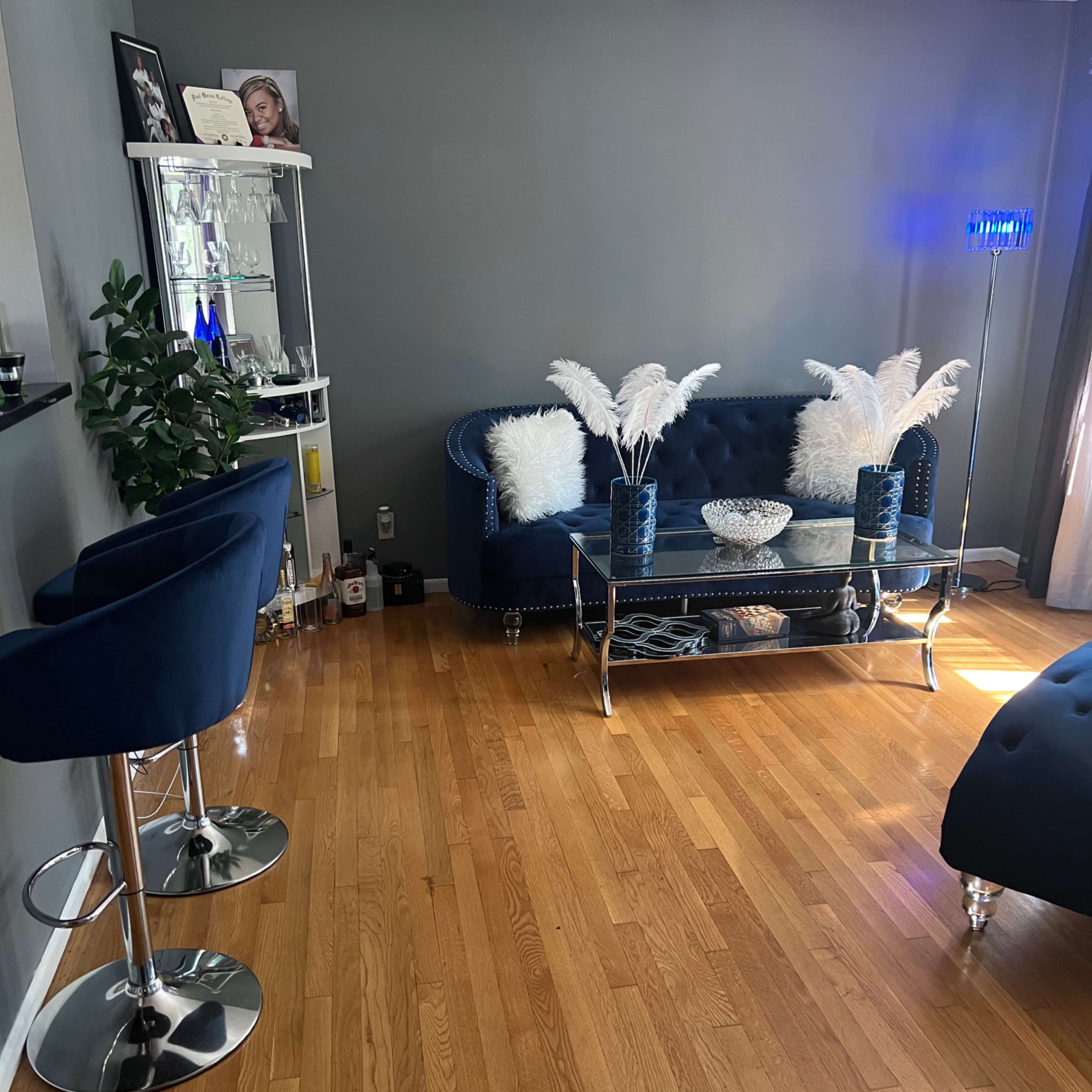 A living room featuring a blue sofa, a glass coffee table with decorative items, and two bar stools on a wooden floor.