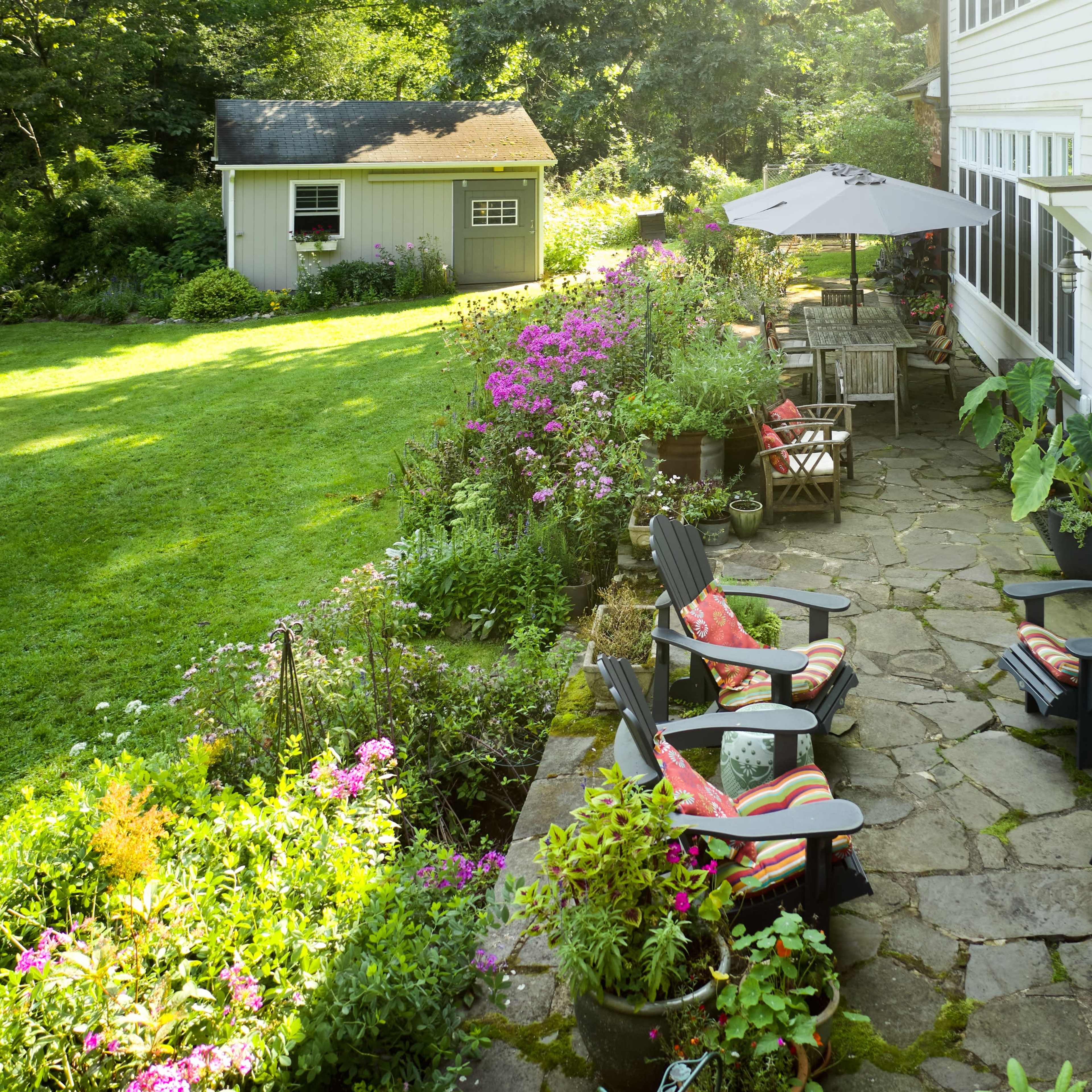 A stone patio with four wooden chairs and colorful cushions overlooks a garden filled with flowers and greenery, alongside a white house and a shed.