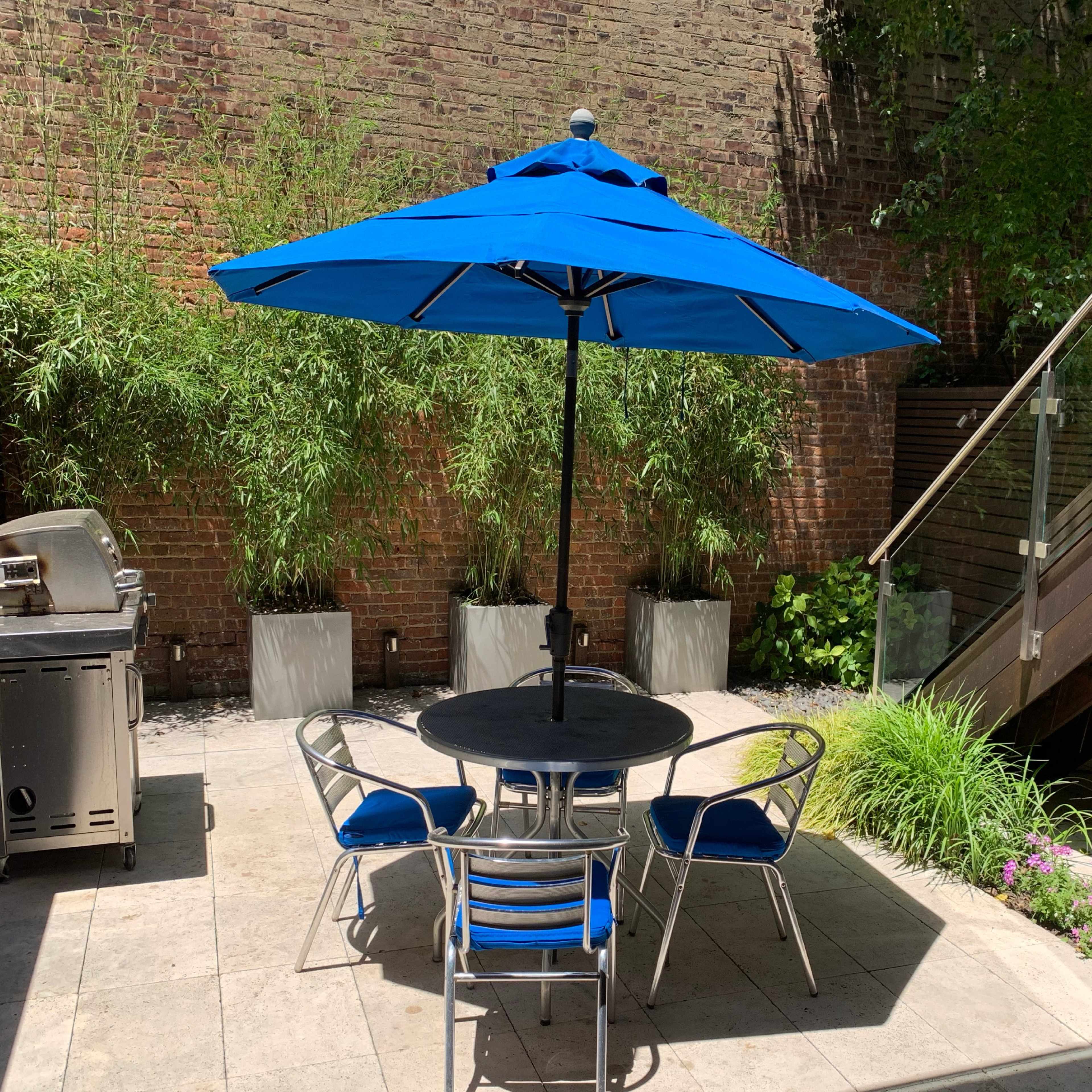 A blue umbrella shades a circular table and four chairs in a patio area surrounded by potted plants and a grill, with a wooden staircase nearby.