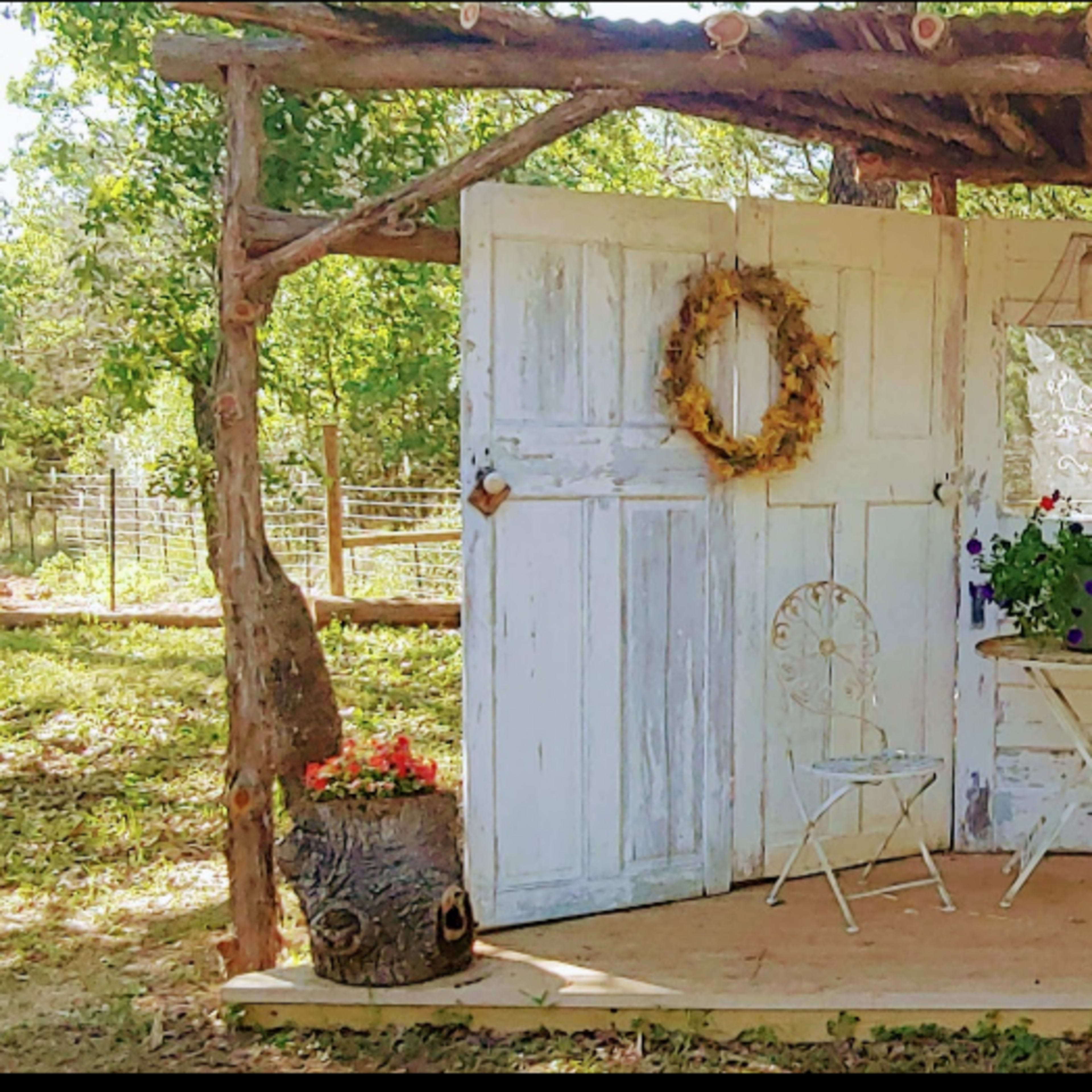 An outdoor setting featuring a rustic wooden structure with multiple white doors, a small round table with a floral arrangement, and two chairs.
