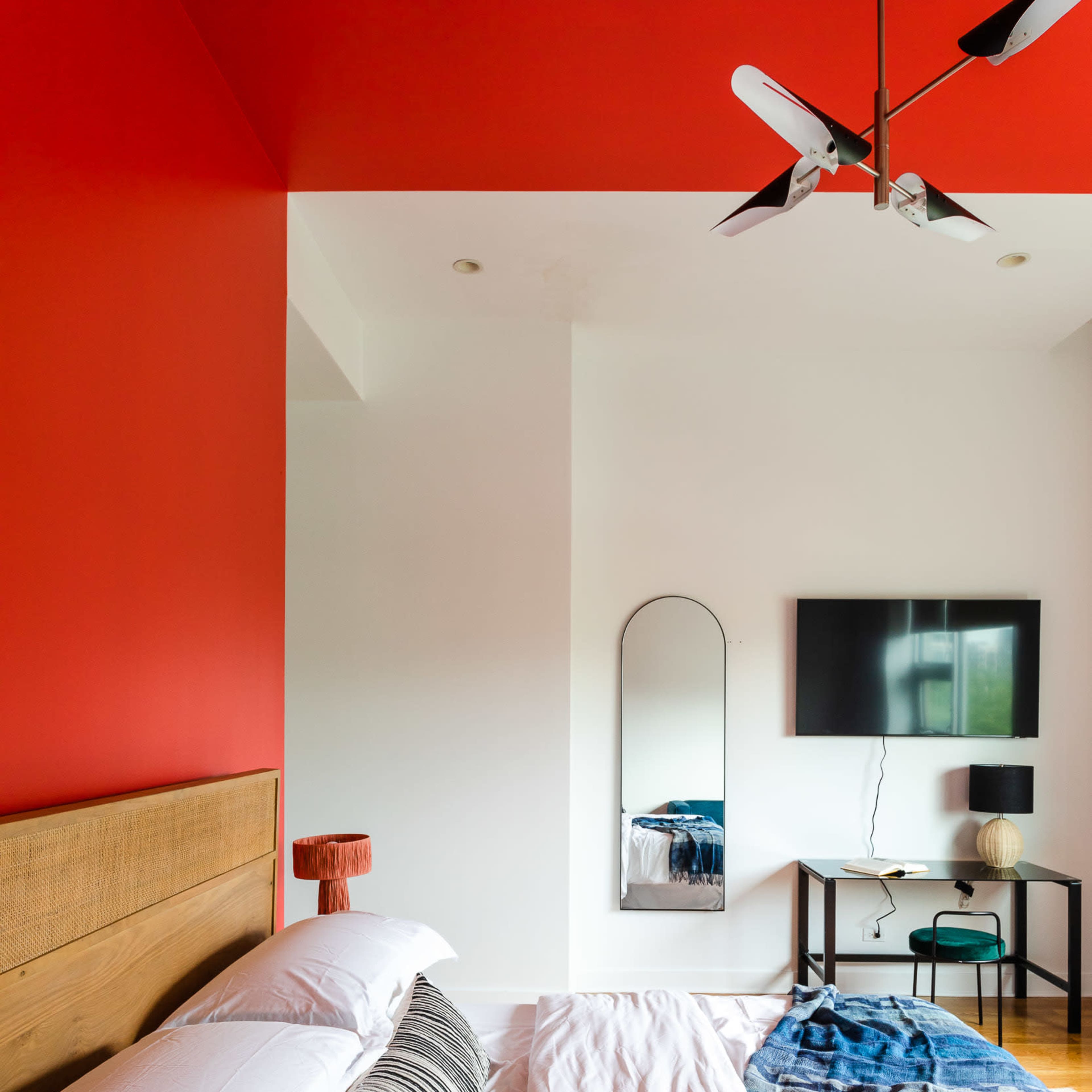 A modern bedroom featuring a red accent wall, a bed with white linens and decorative pillows, a flat-screen TV mounted on the wall, and a small table with a lamp.