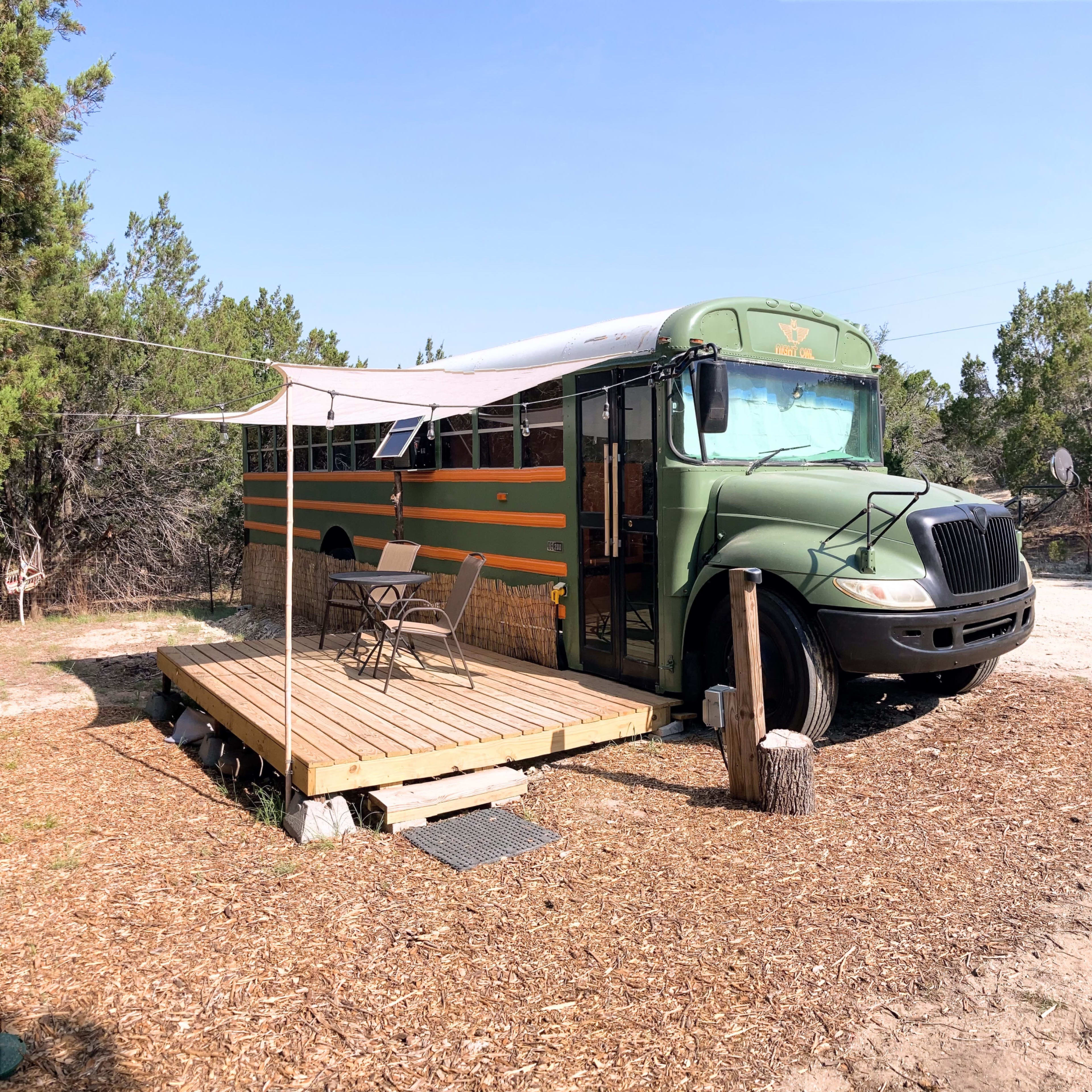 A converted bus is parked next to a wooden deck surrounded by trees in a rural setting.