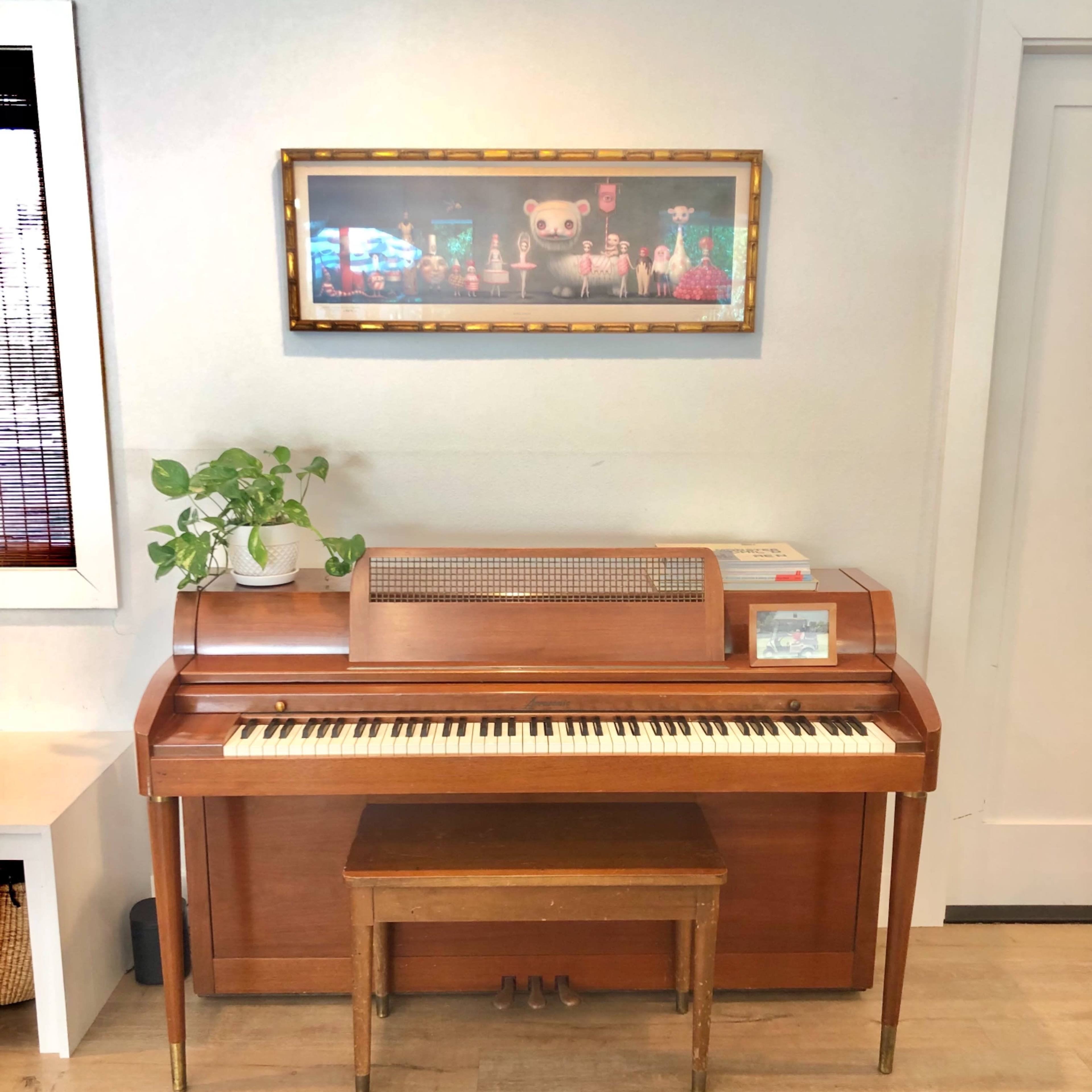 A wooden piano with a small bench is positioned against a wall, adorned with a framed artwork above it and a potted plant nearby.